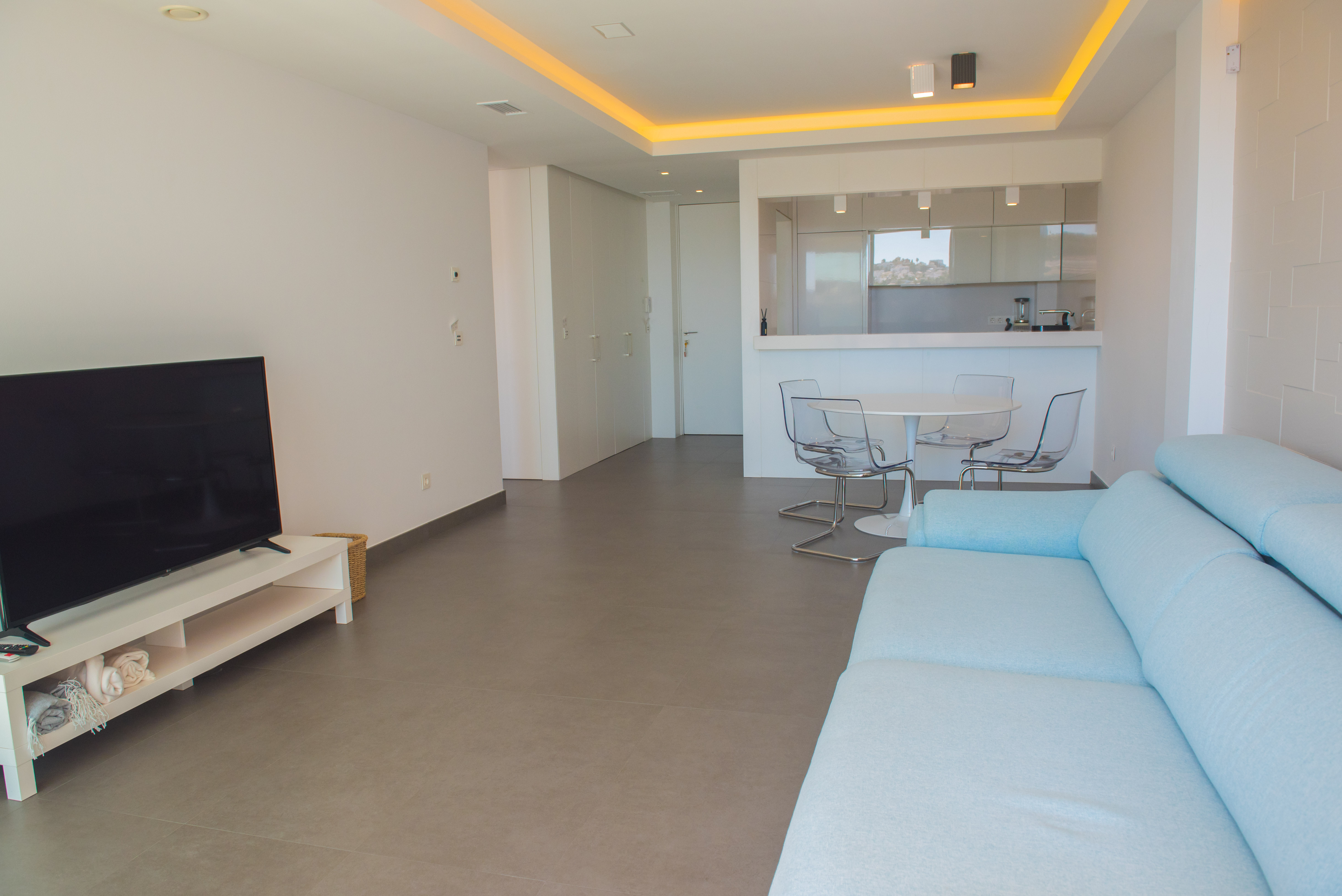 Open woonkamer met lichtblauwe bank, eethoek met transparante stoelen en een geïntegreerde keuken met indirecte plafondverlichting.