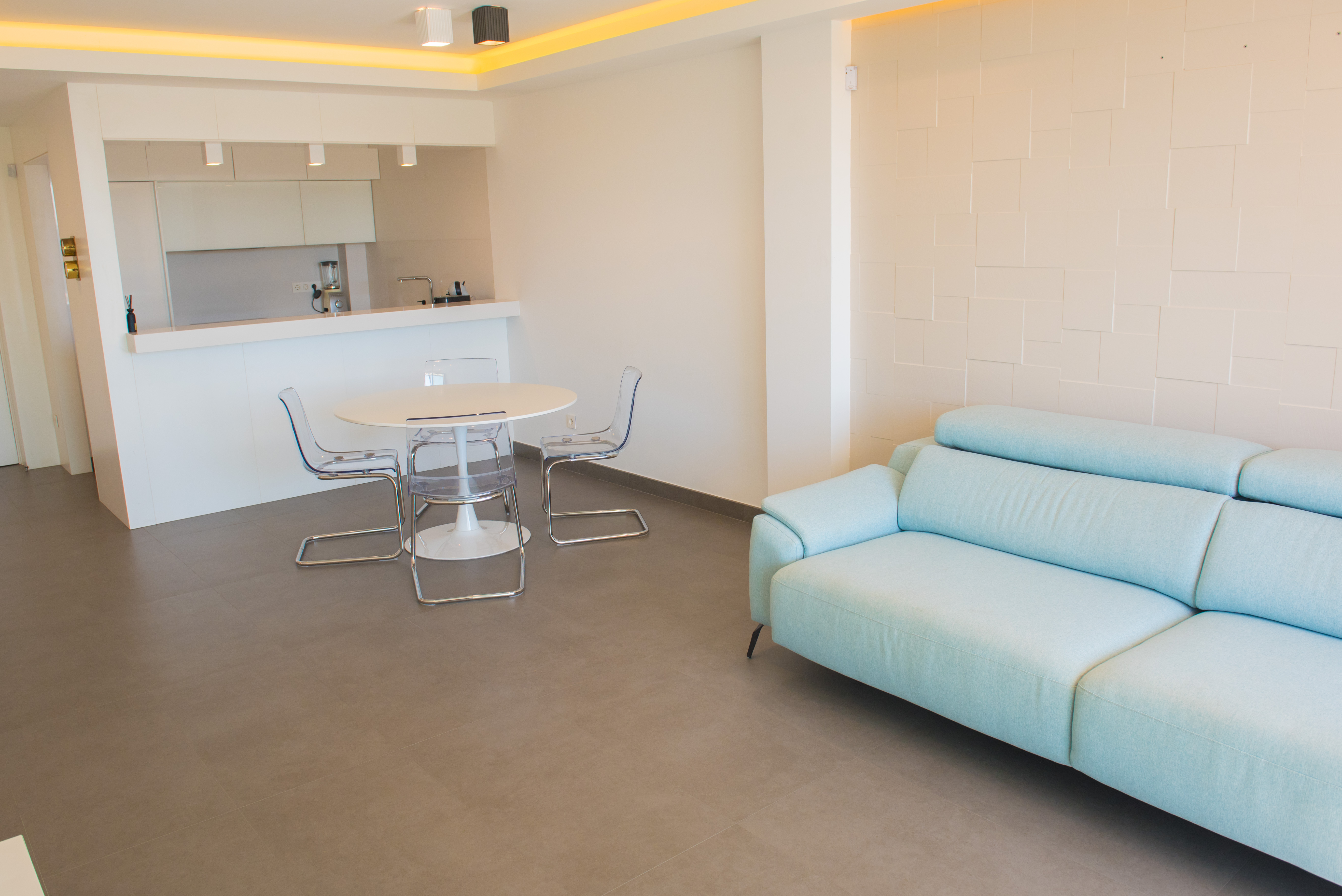 Open woonruimte met lichtblauwe bank, transparante stoelen en een witte eettafel. Voorzien van een wand met geometrisch reliëf en indirecte plafondverlichting.