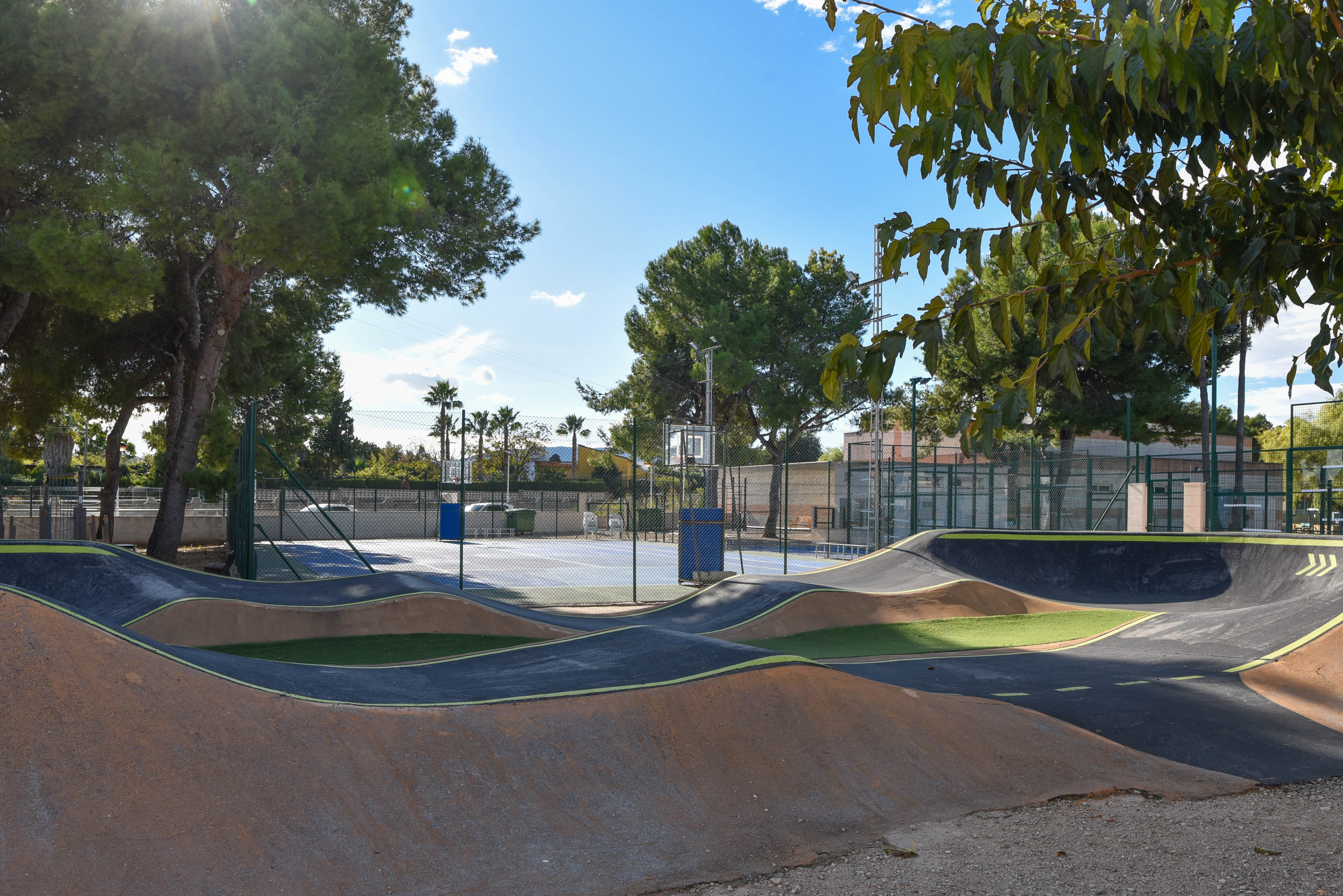 Geasfalteerde pump track met golvende banen naast een omheind multisportveld, omgeven door volgroeide pijnbomen in een recreatiegebied.