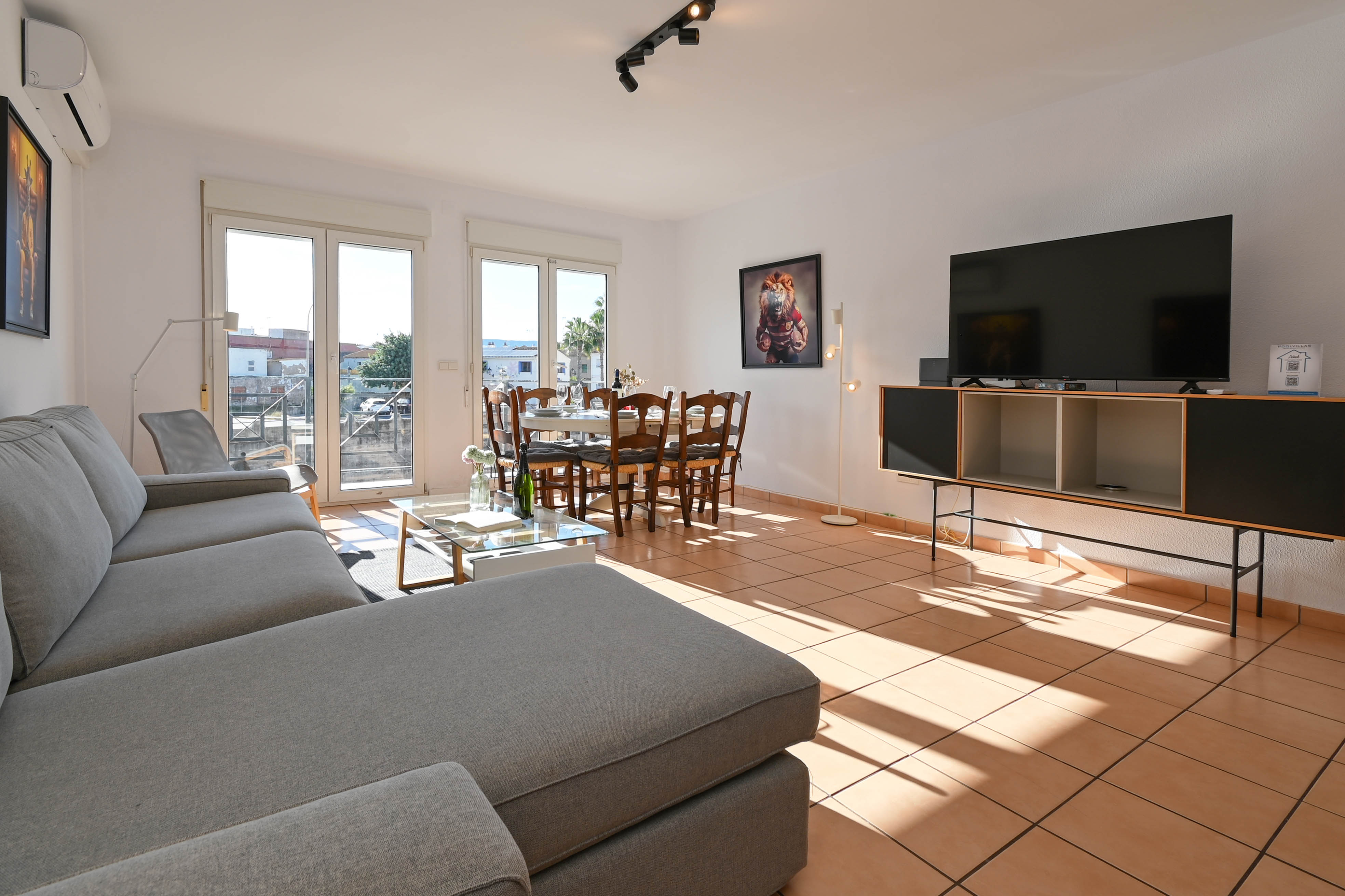 Lichte open woon- en eetkamer met terracotta tegelvloer, een grote grijze hoekbank en directe toegang tot het balkon via glazen schuifdeuren.