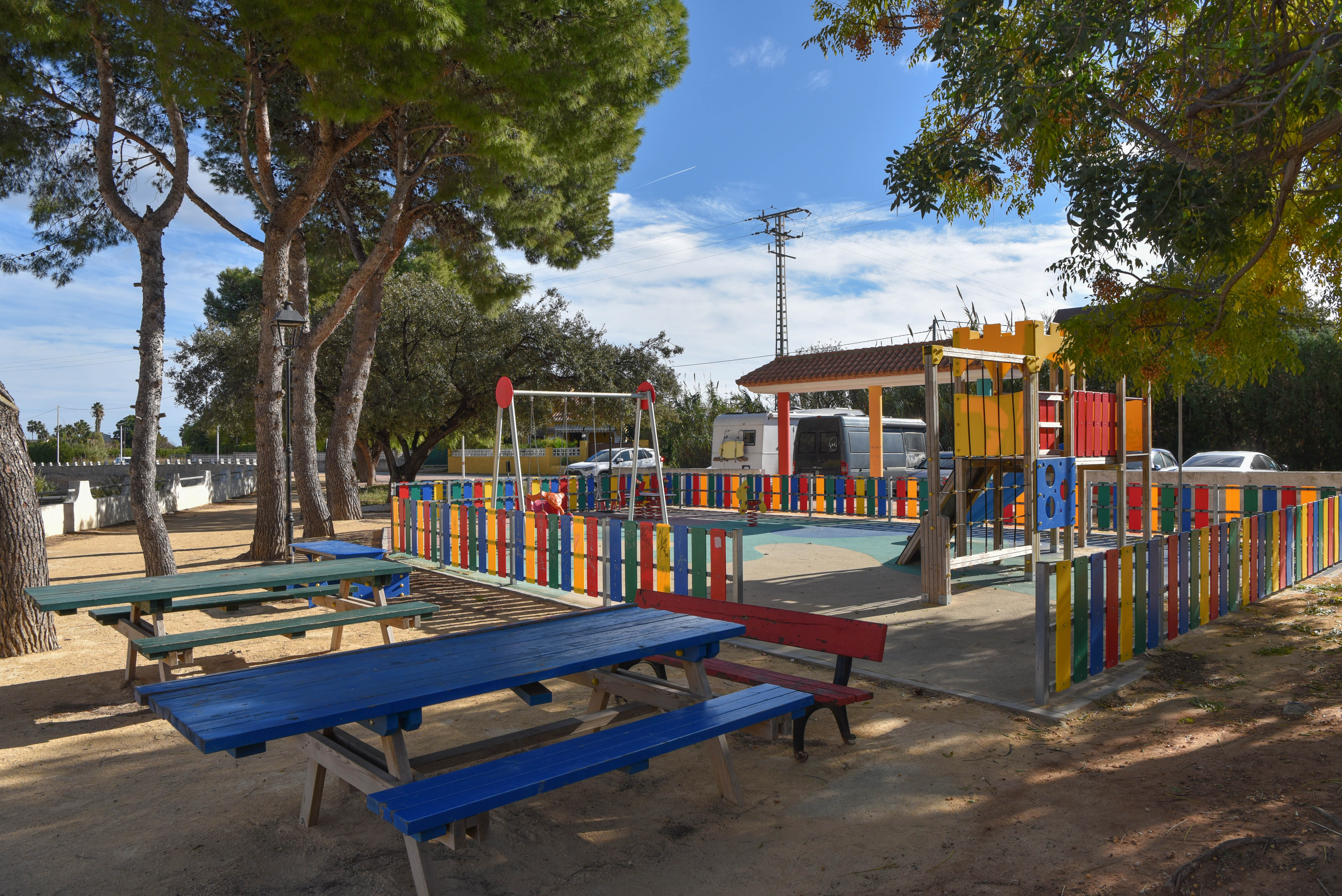 Omheinde kinderspeelplaats met kleurrijke toestellen en houten picknicktafels onder de schaduw van volwassen pijnbomen in een zonnige omgeving.