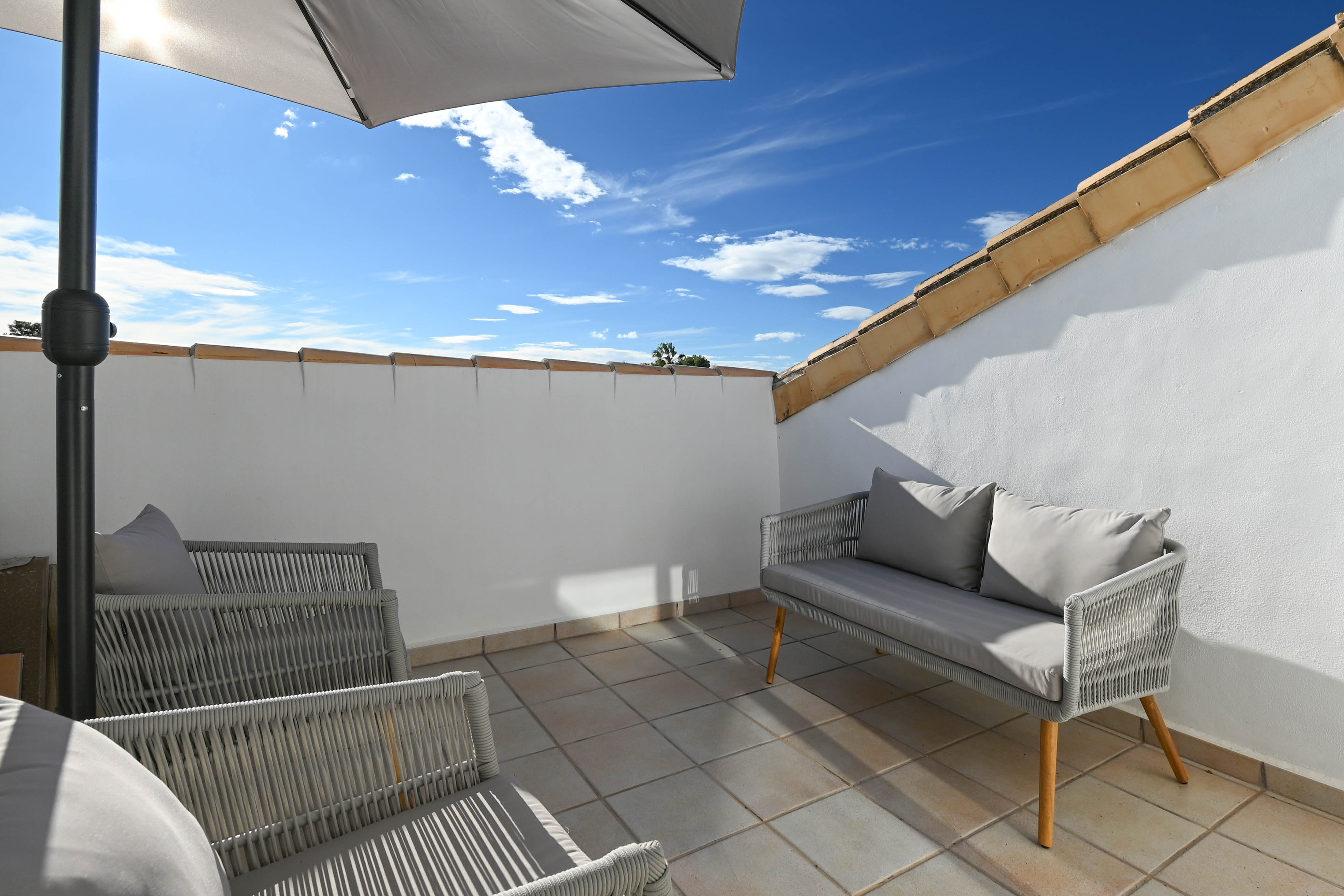 Privé dakterras met grijs gevlochten buitenmeubilair, tegelvloer en parasol, gelegen onder de blauwe lucht van de Costa Blanca.
