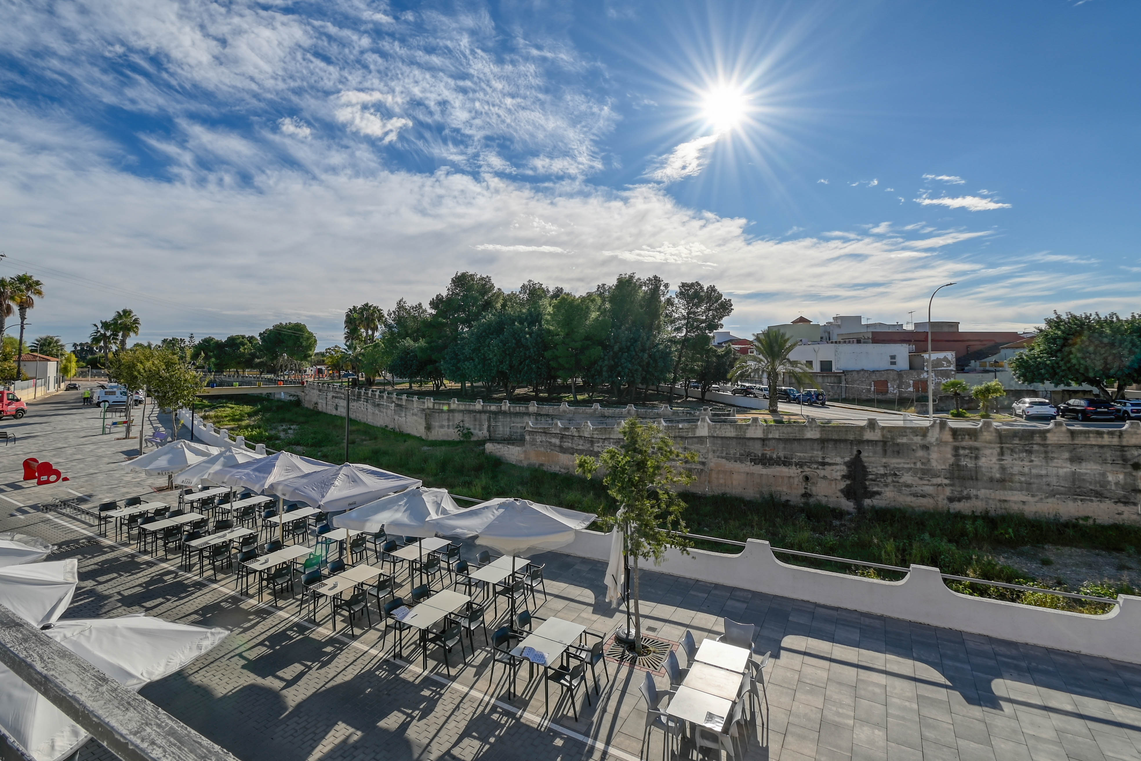 Verhoogd terras met witte parasols en zitgelegenheid, uitkijkend over een stenen kanaalmuur en een groene omgeving onder een stralende zon.