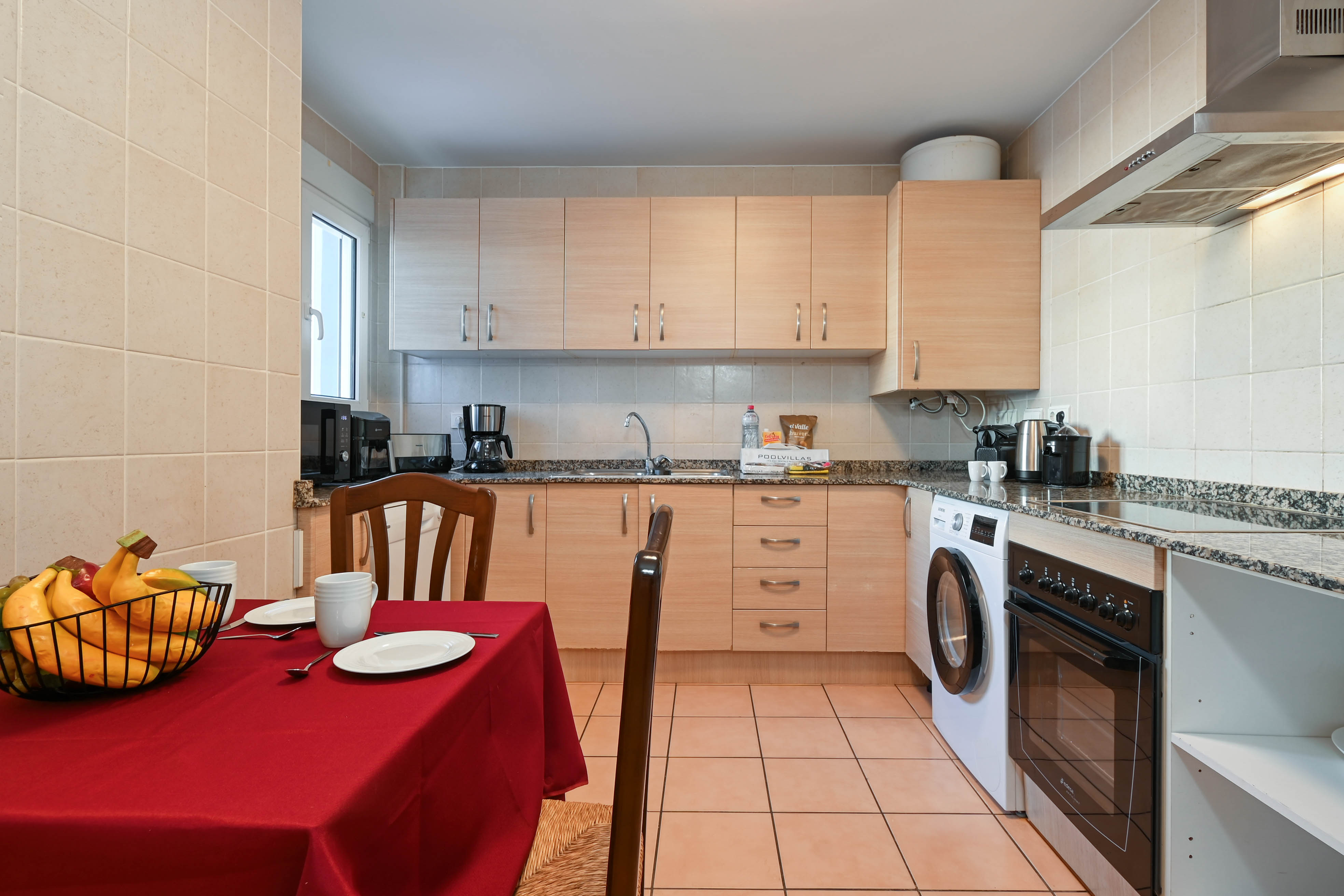 Volledig uitgeruste keuken met licht houten kasten, granieten werkbladen en inbouwapparatuur, inclusief een eethoek met rood tafelkleed.