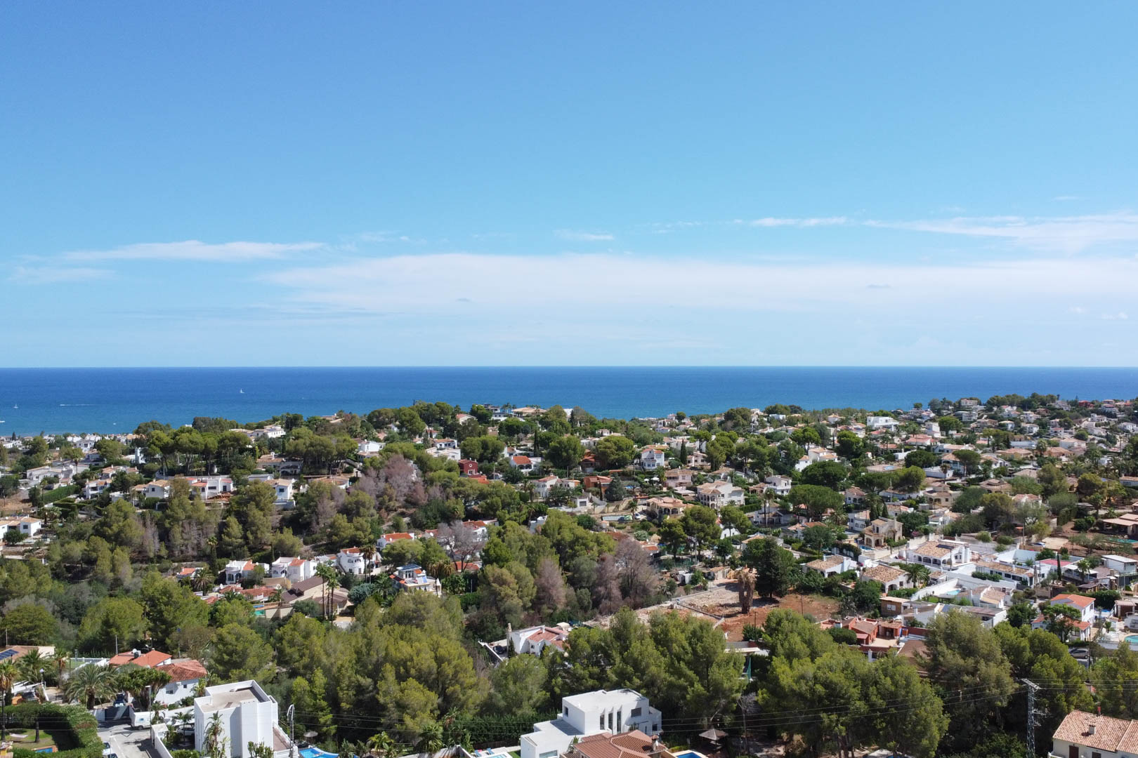 Panoramisch luchtfoto van een residentiële wijk in Dénia, met witte villa's tussen mediterrane pijnbomen en een onbelemmerd uitzicht op de Middellandse Zee.