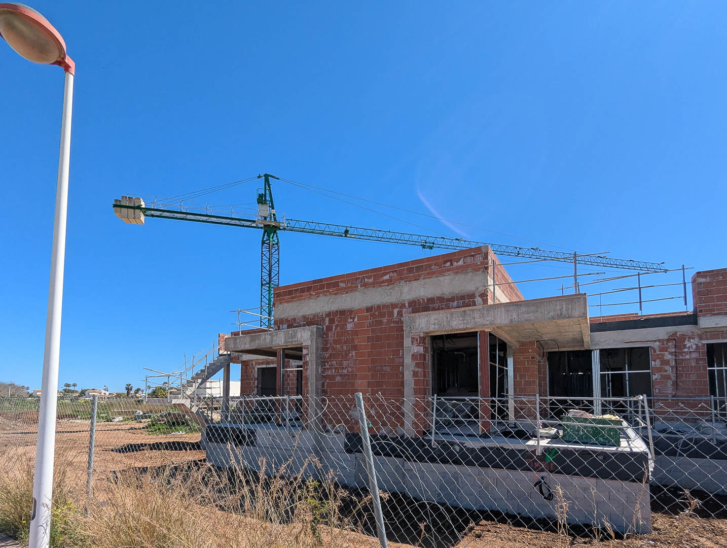 Huidige bouwfase van de villa met de bakstenen en betonnen ruwbouw, voorzien van een bouwkraan en omheining onder een onbewolkte blauwe hemel.