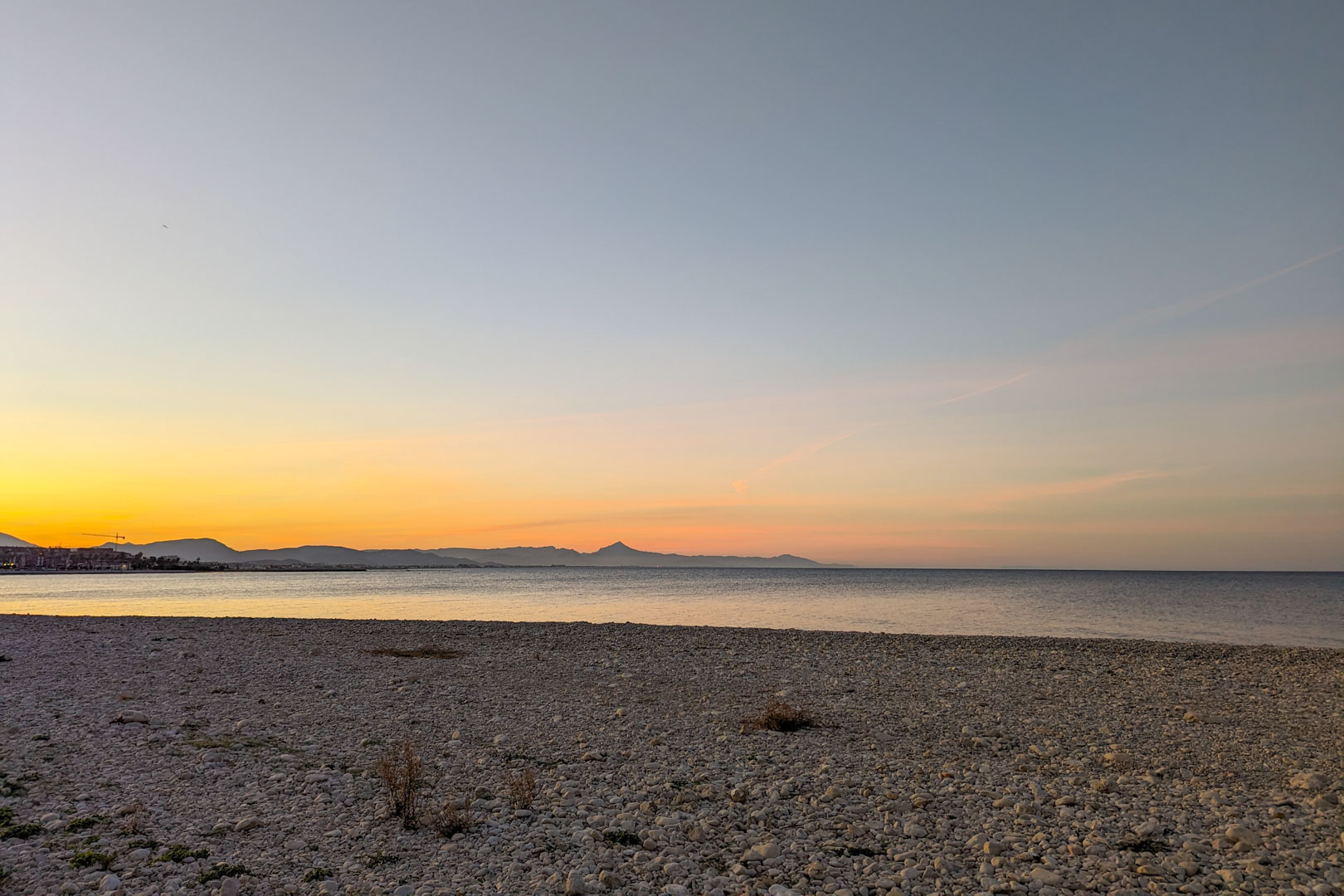 Panoramisch uitzicht op het nabijgelegen kiezelstrand bij zonsondergang, met bergsilhouetten aan de horizon en een kalme Middellandse Zee.