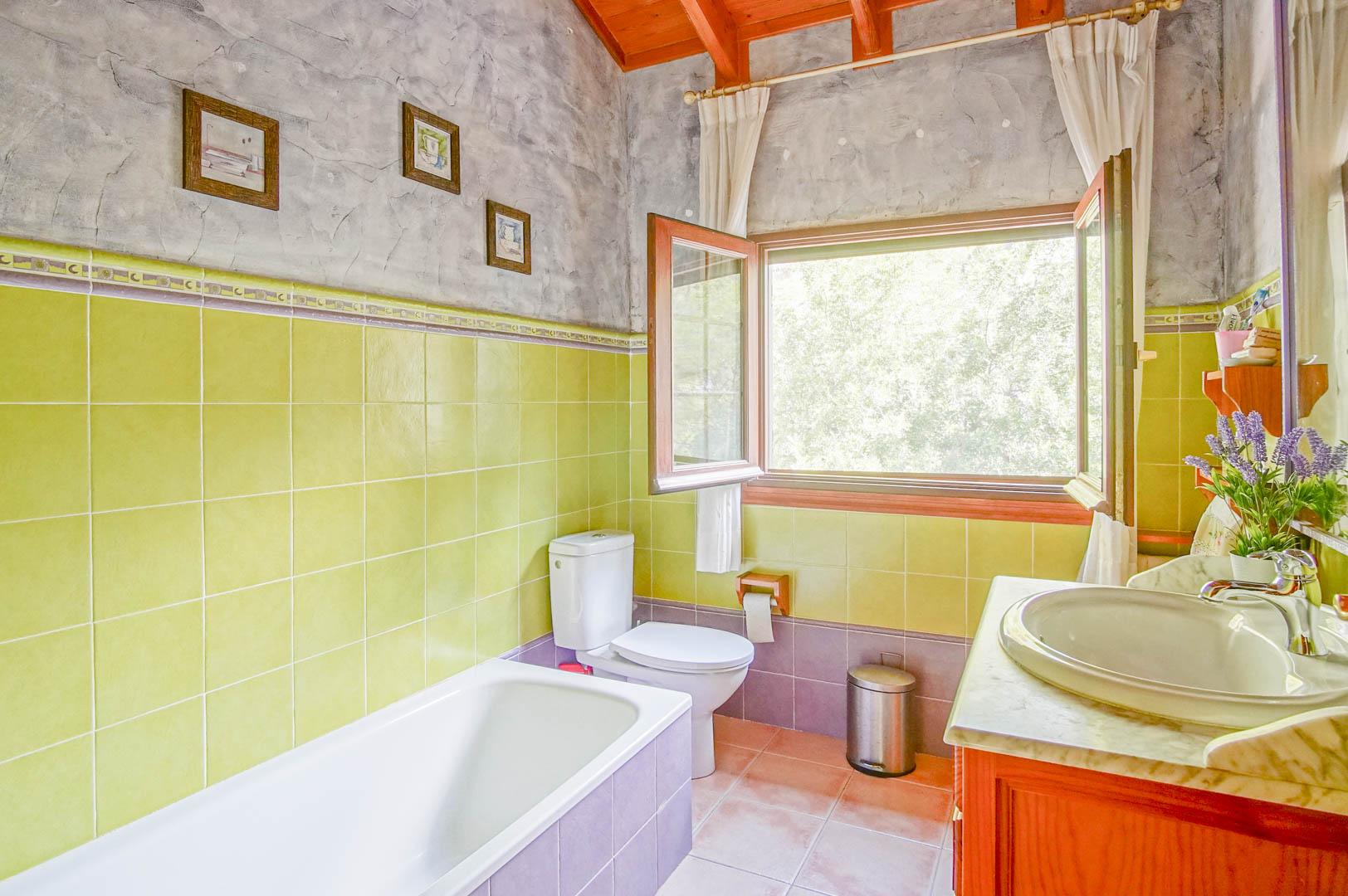 Badkamer met houten balkenplafond, groene betegeling en een marmeren wastafelblad. Een groot raam biedt natuurlijk licht en uitzicht op de groene omgeving.