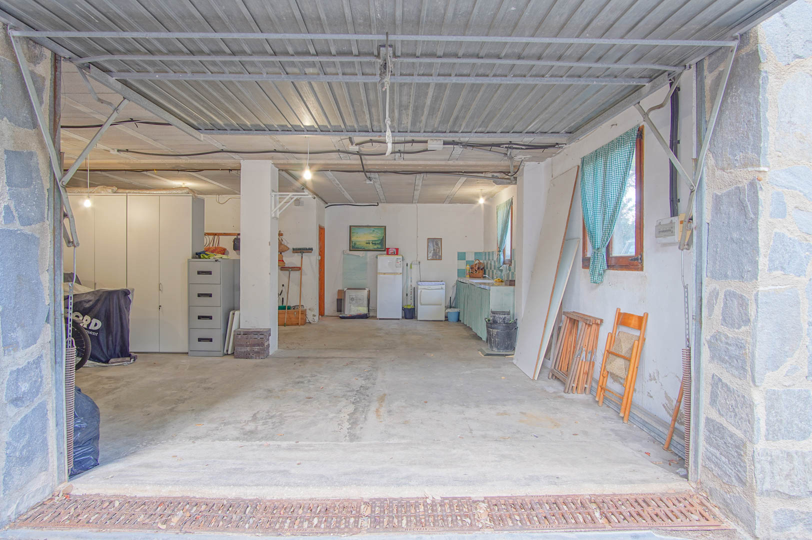 Ruime halfondergrondse verdieping met garage voor meerdere auto's, werkplaats en kitchenette. Voorzien van betonnen vloeren, stenen pilaren en een metalen kanteldeur.