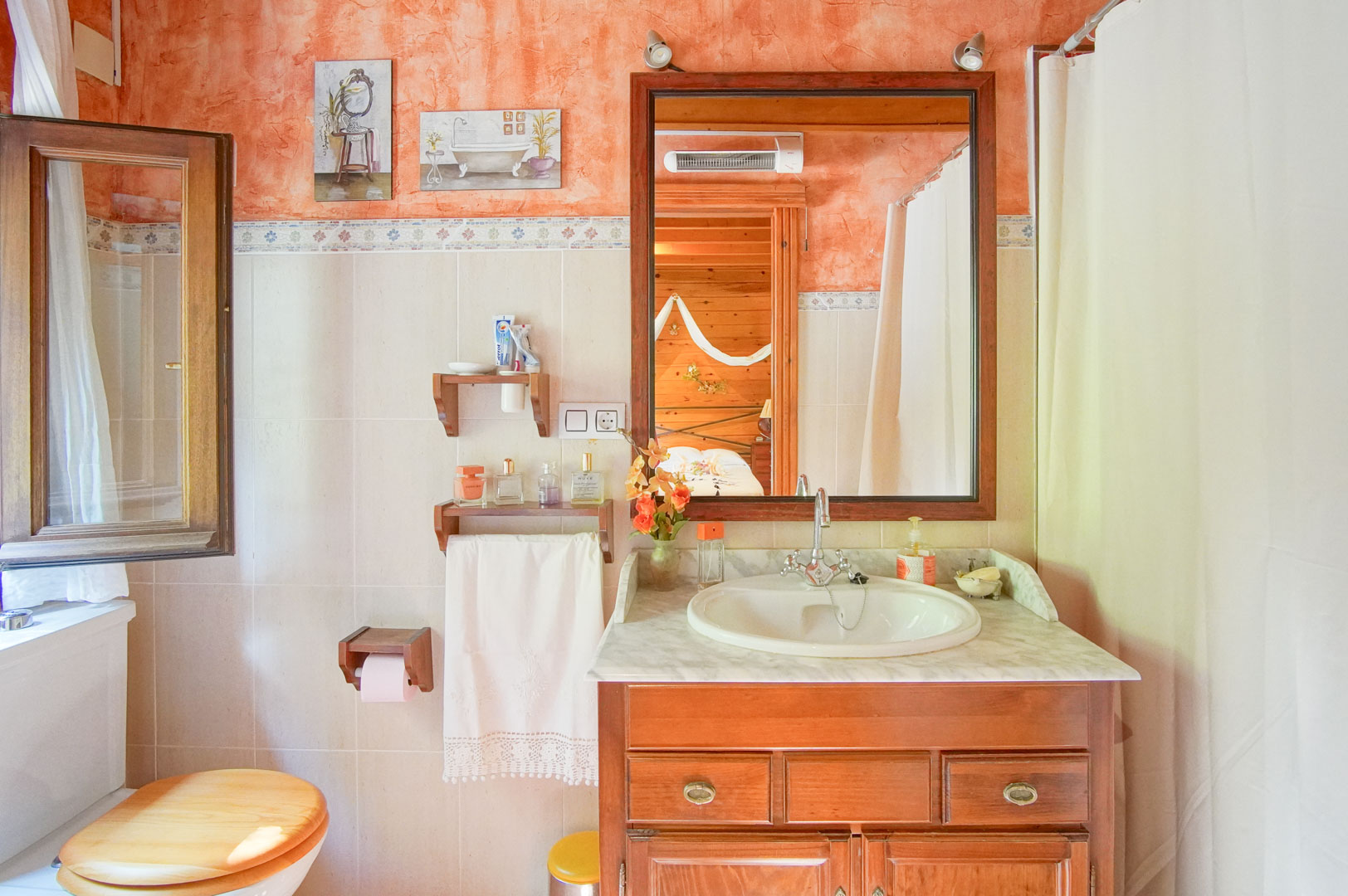 Rustieke badkamer met houten wastafelmeubel, marmeren blad en terracotta wanden, met in de spiegel een doorkijk naar de slaapkamer met grenen houten afwerking.