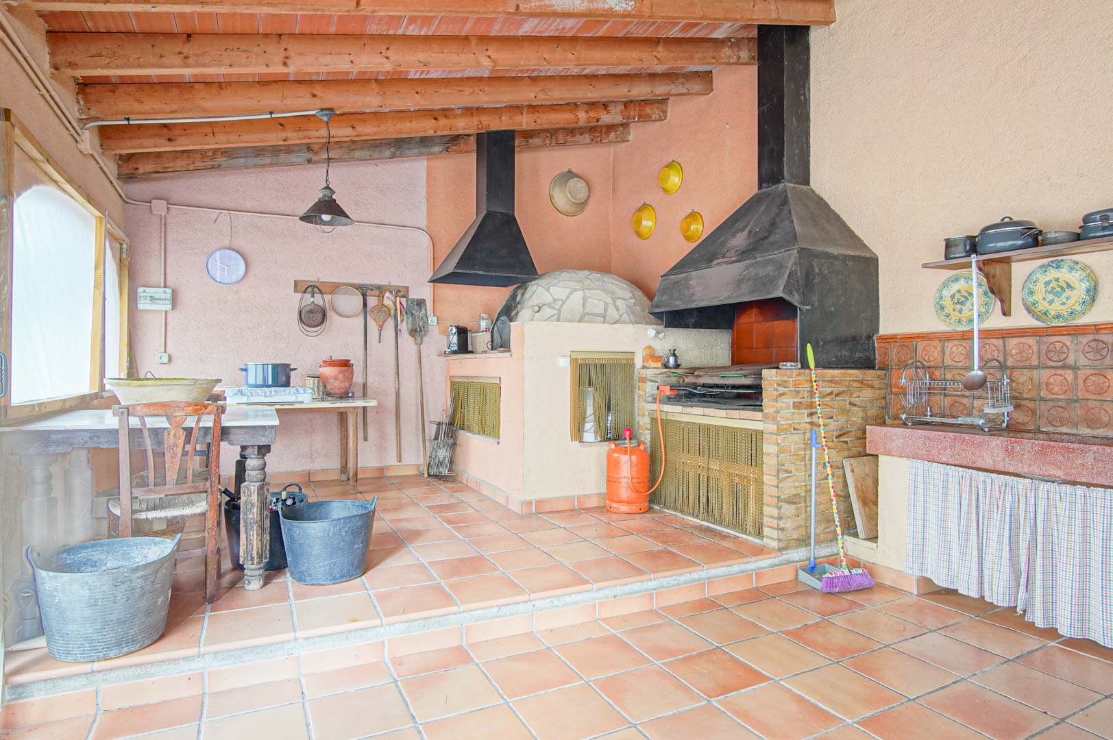 Rustieke buitenkeuken met een traditionele houtoven, gemetselde barbecue en een plafond met houten balken en terracotta vloer.