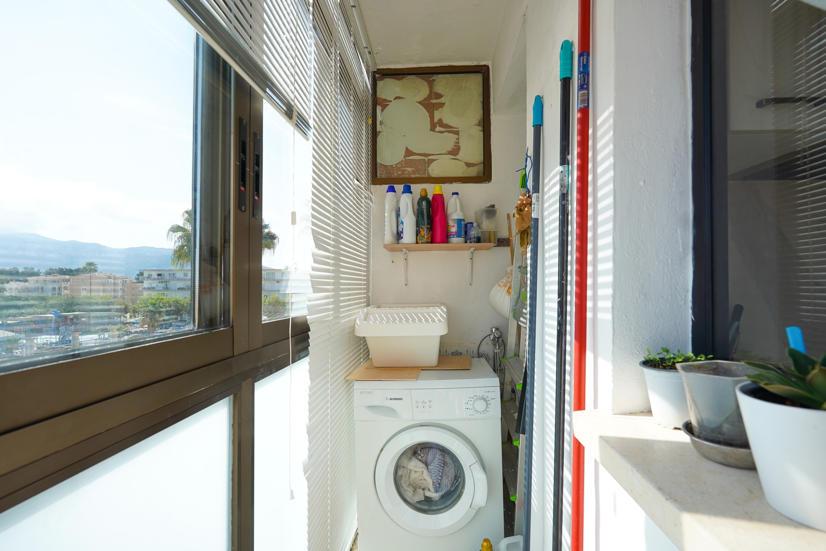 Afgesloten wasruimte op het balkon met een voorlader wasmachine, opbergschap voor schoonmaakmiddelen en grote ramen met horizontale jaloezieën.