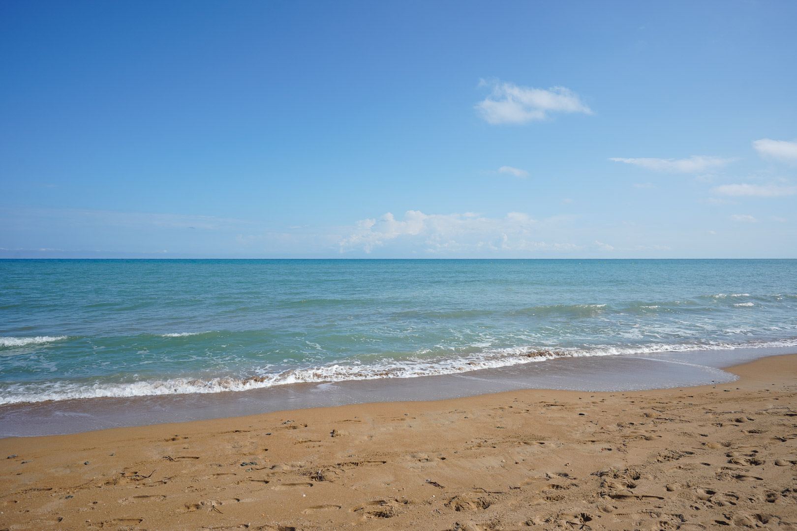 Breed zandstrand met zachte turquoise golven onder een helderblauwe lucht. De kustlijn toont goudkleurig zand en kalm mediterraan water.