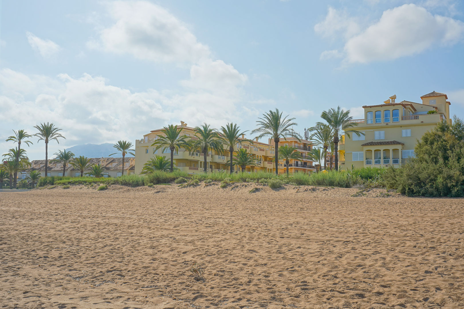 Breed zandstrand voor woongebouwen in mediterrane stijl met palmbomen en uitzicht op de bergen in de verte onder een heldere hemel.