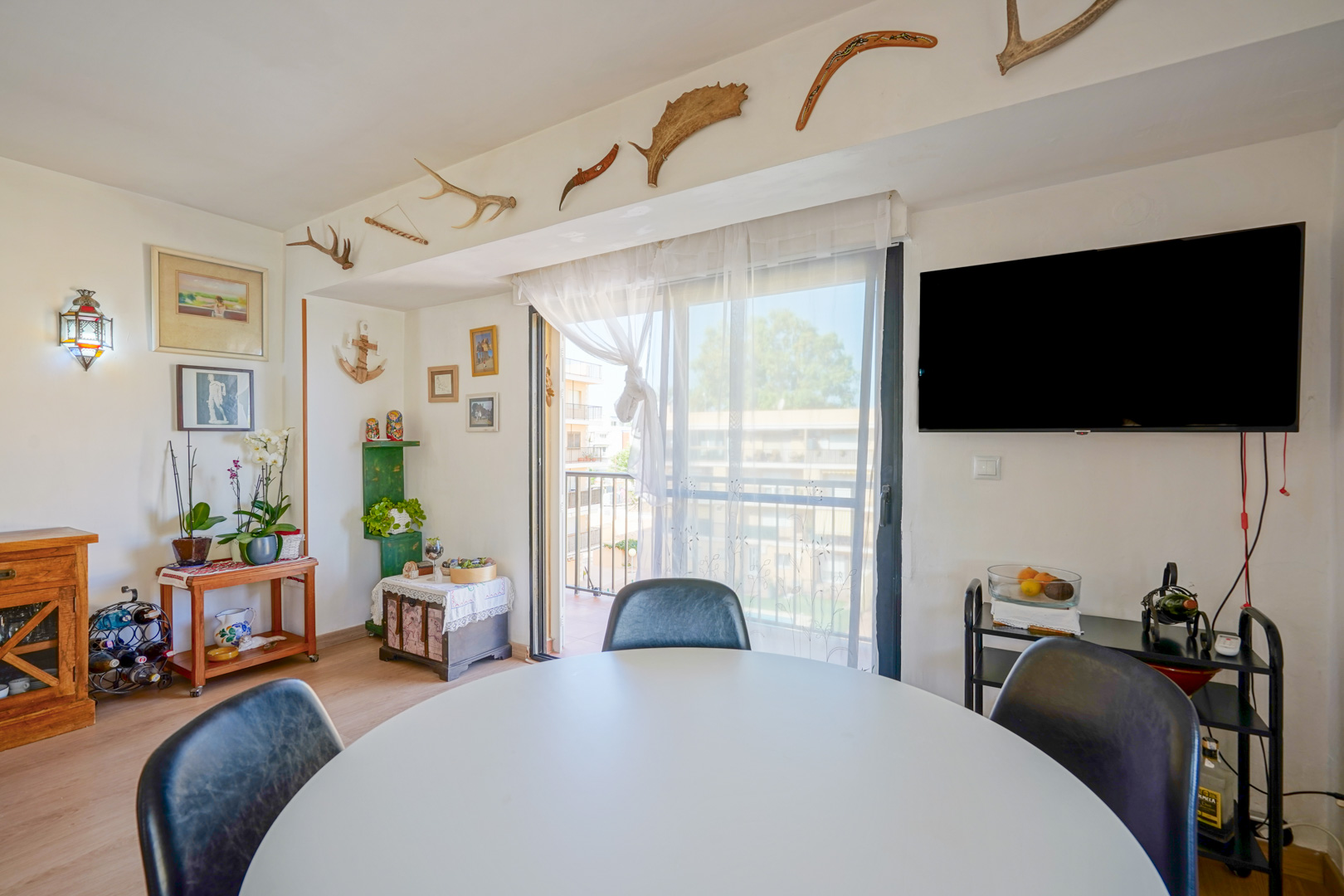 Eethoek met een witte ronde tafel, wandtelevisie en toegang tot het balkon. De wand is uniek gedecoreerd met geweien en boemerangs boven de schuifpui.