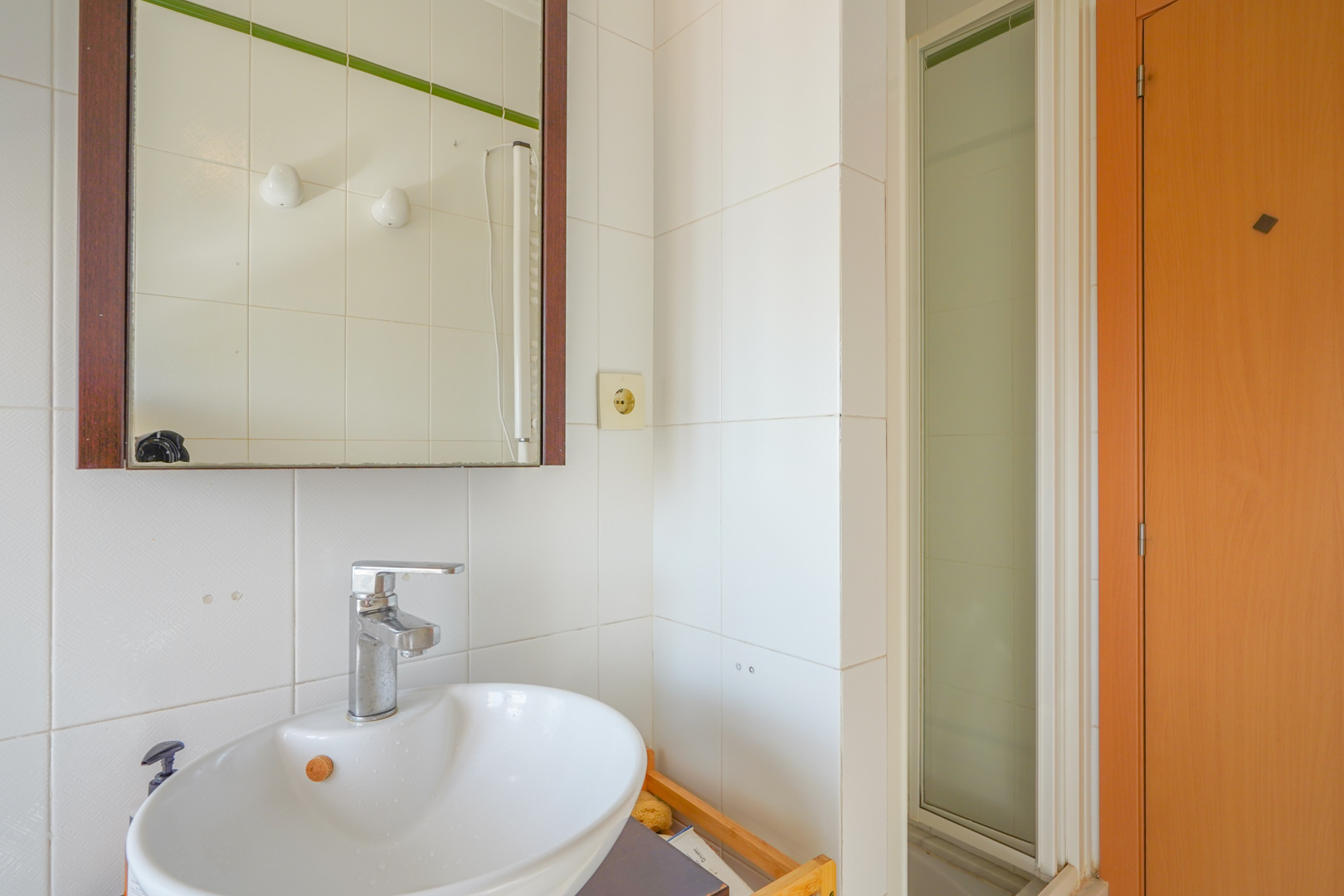 Functionele badkamer met witte keramische wastafel, chromen kraan en spiegel met houten omlijsting, naast een douchecabine met matglas.