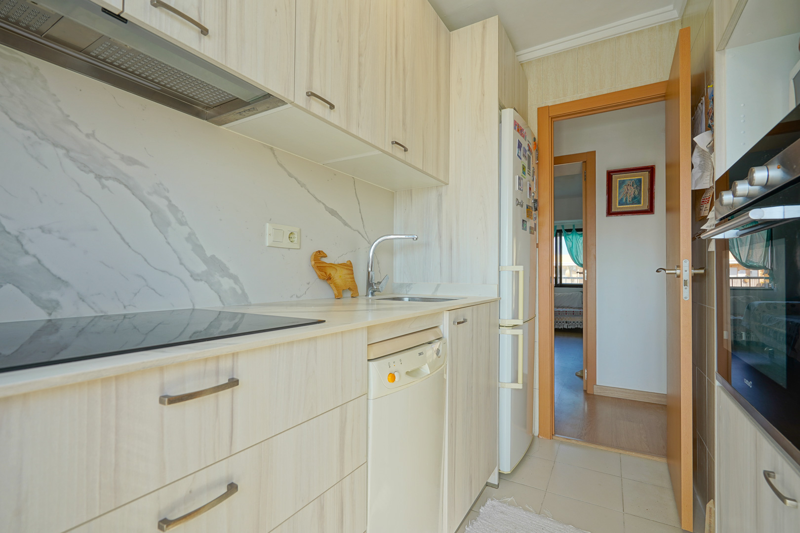 Functionele keuken met lichte houten kasten, marmerlook achterwand, inductiekookplaat en inbouwapparatuur met doorkijk naar de gang.