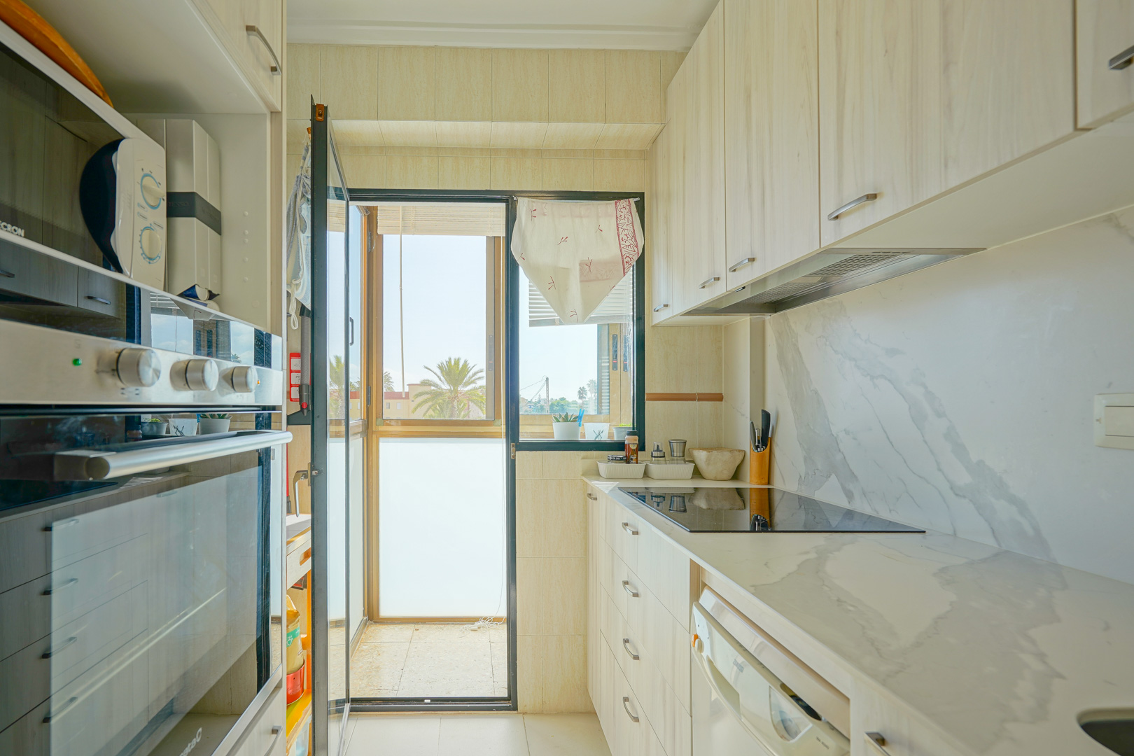 Keuken met lichte houten kasten, aanrechtblad met marmerlook en een glazen deur naar een balkon met uitzicht op palmbomen.