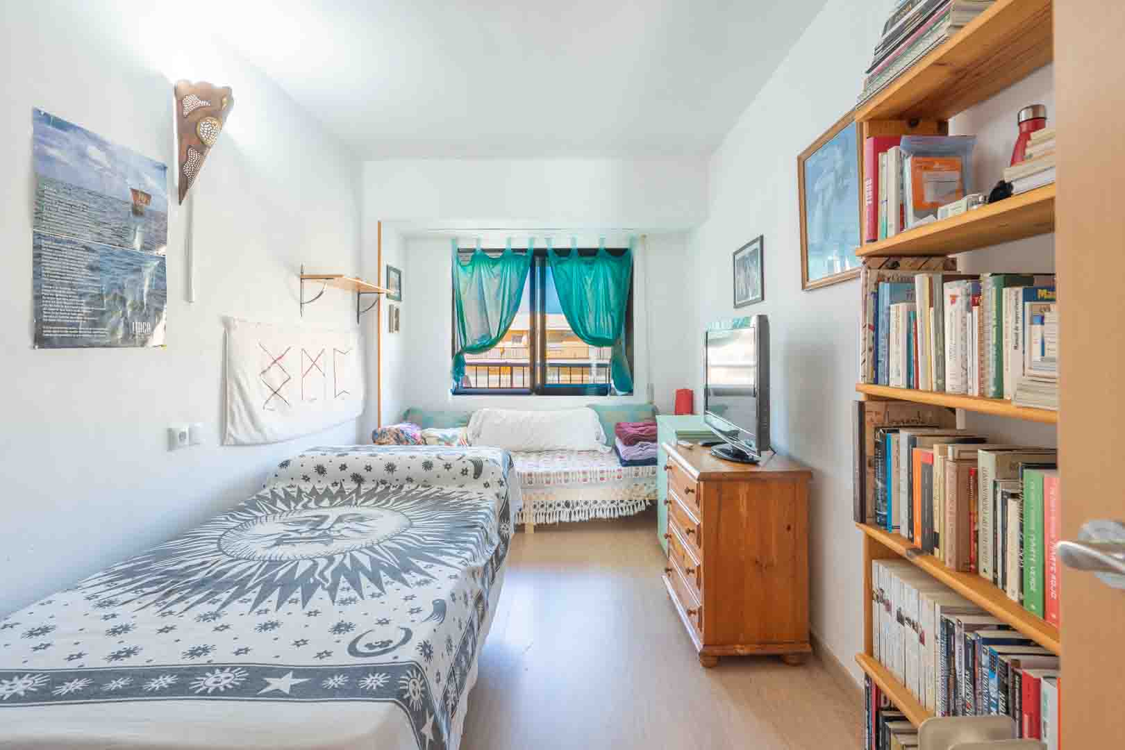 Lichte slaapkamer met een eenpersoonsbed, houten boekenkast en ladekast met televisie. Natuurlijk licht valt binnen via een raam met turquoise gordijnen.