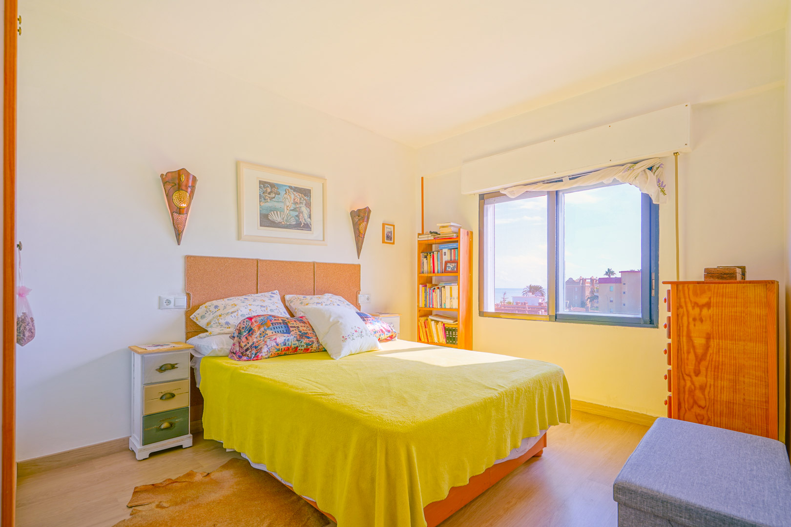 Lichte slaapkamer met kurken hoofdbord, gele sprei en houten meubels. Het grote raam biedt natuurlijk licht en uitzicht op de omliggende bebouwing.