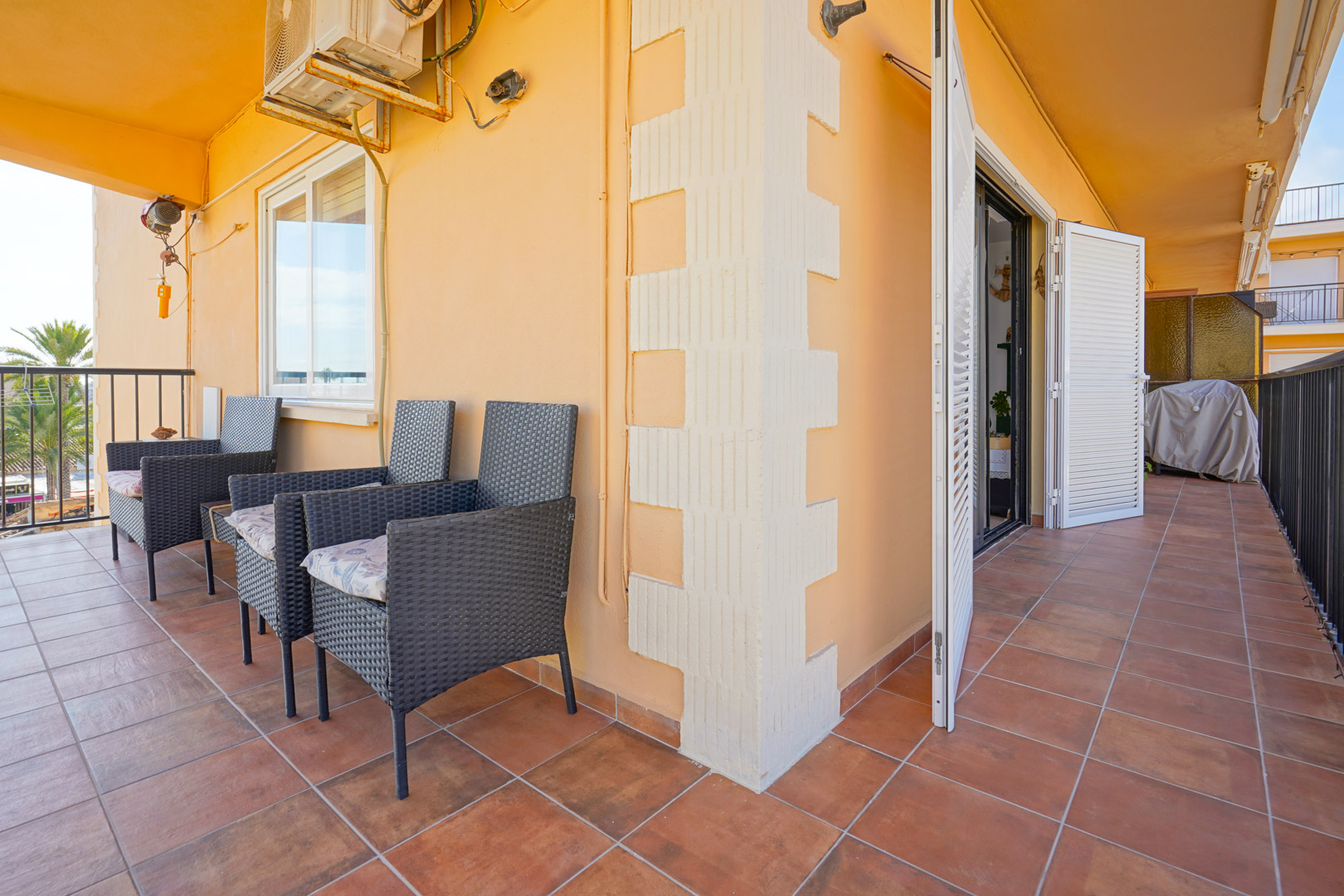 Overdekt terras met terracotta vloertegels, donkere wicker fauteuils en witte luiken. De buitenruimte is voorzien van een airconditioningunit en metalen hekwerk.