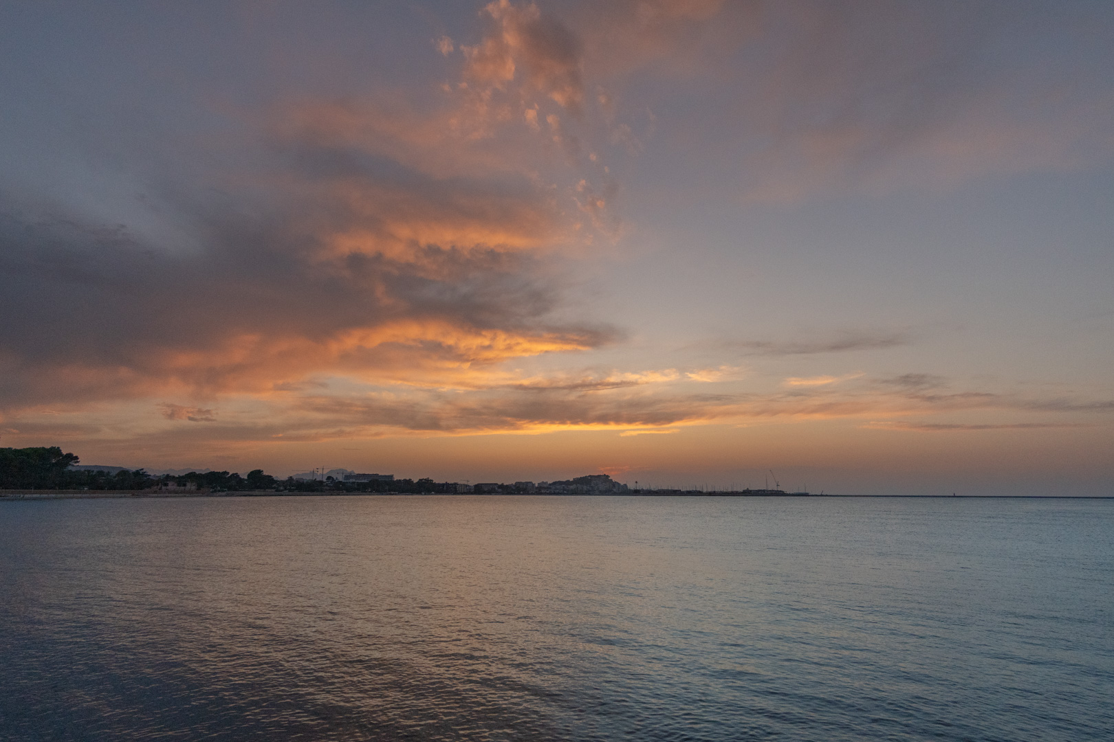 Panoramisch uitzicht op de Middellandse Zeekust bij zonsondergang, met oranje getinte wolken en kalm water dat de avondlucht weerspiegelt boven de verre stad.
