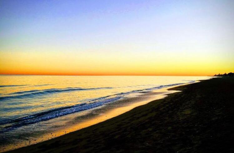 Panoramisch uitzicht op een zandstrand bij zonsondergang, met kabbelende golven en een kleurovergang van diepblauw naar goudgeel aan de horizon.