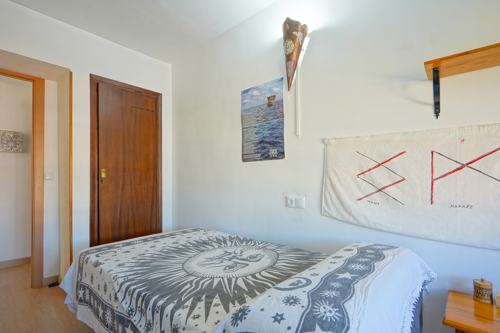 Slaapkamer met eenpersoonsbed en sprei met zonmotief, witte wanden met een koperen wandlamp en een massieve donkerhouten deur.