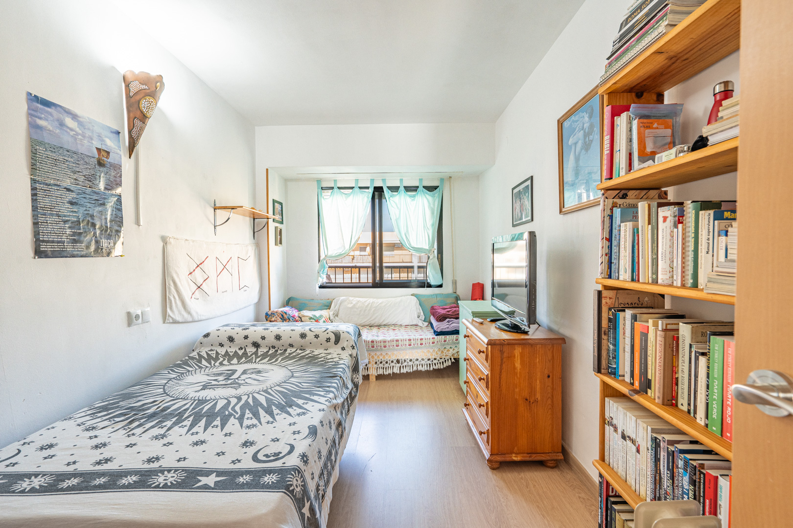 Slaapkamer met eenpersoonsbed, houten boekenkast en ladekast. Voorzien van laminaatvloer en een raam met lichtblauwe gordijnen voor natuurlijk licht.