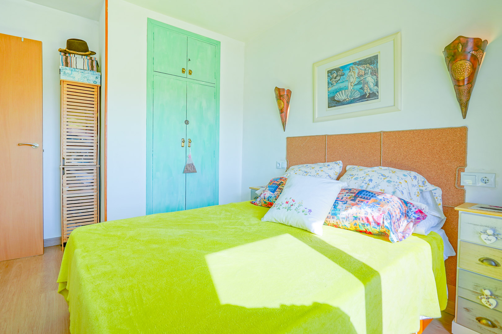 Slaapkamer met kurken hoofdbord, mintgroene inbouwkast en klassiek kunstwerk. Natuurlijk licht verlicht de limoengroene sprei en het rustieke nachtkastje.