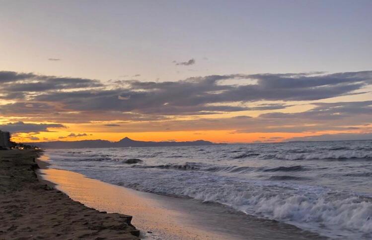 Uitzicht op de zonsondergang over een zandstrand met brekende golven en bergsilhouetten onder een bewolkte oranje hemel.
