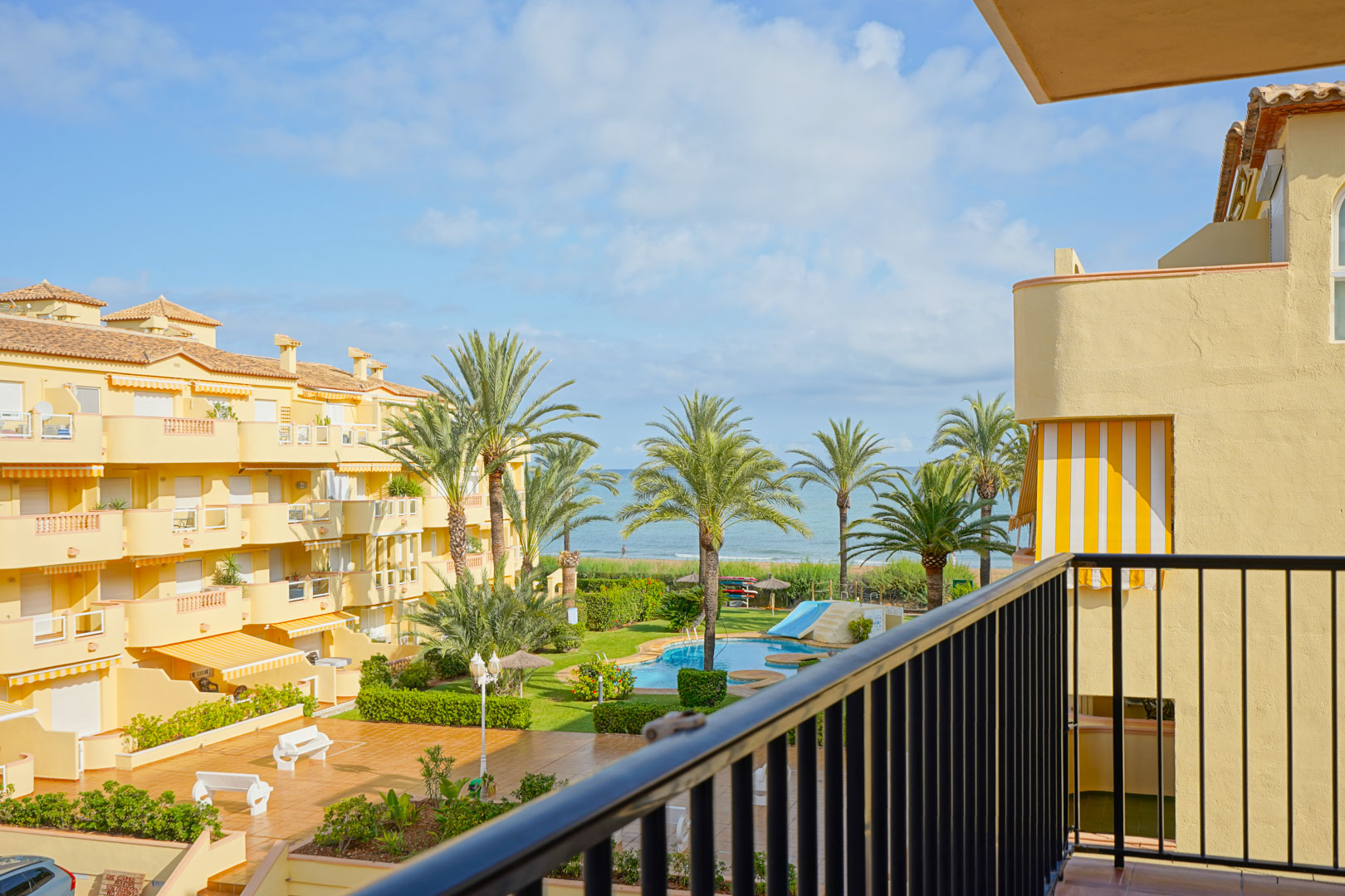 Uitzicht vanaf een privébalkon op een residentieel complex met zwembad, palmbomen en een weids panorama over de Middellandse Zee.