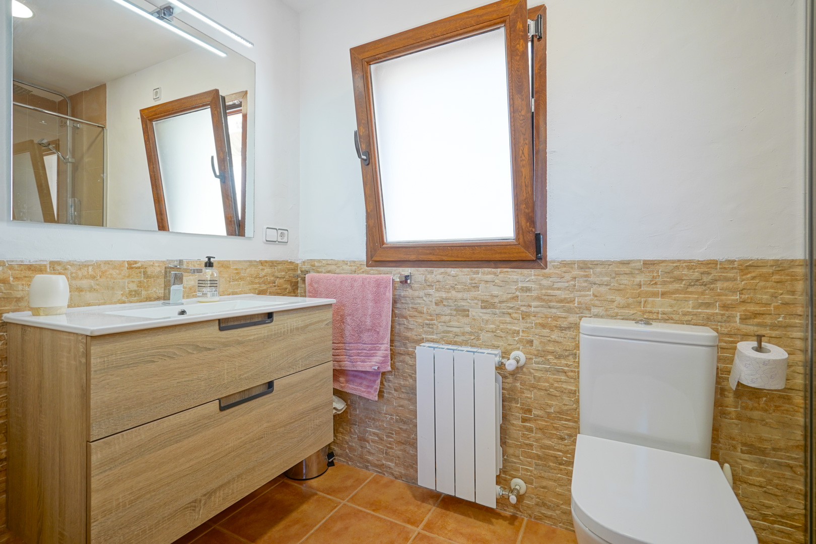 Badkamer met houten wastafelmeubel, LED-verlichte spiegel en wandtegels met steeneffect. Voorzien van een radiator en een houten draaikiepraam.