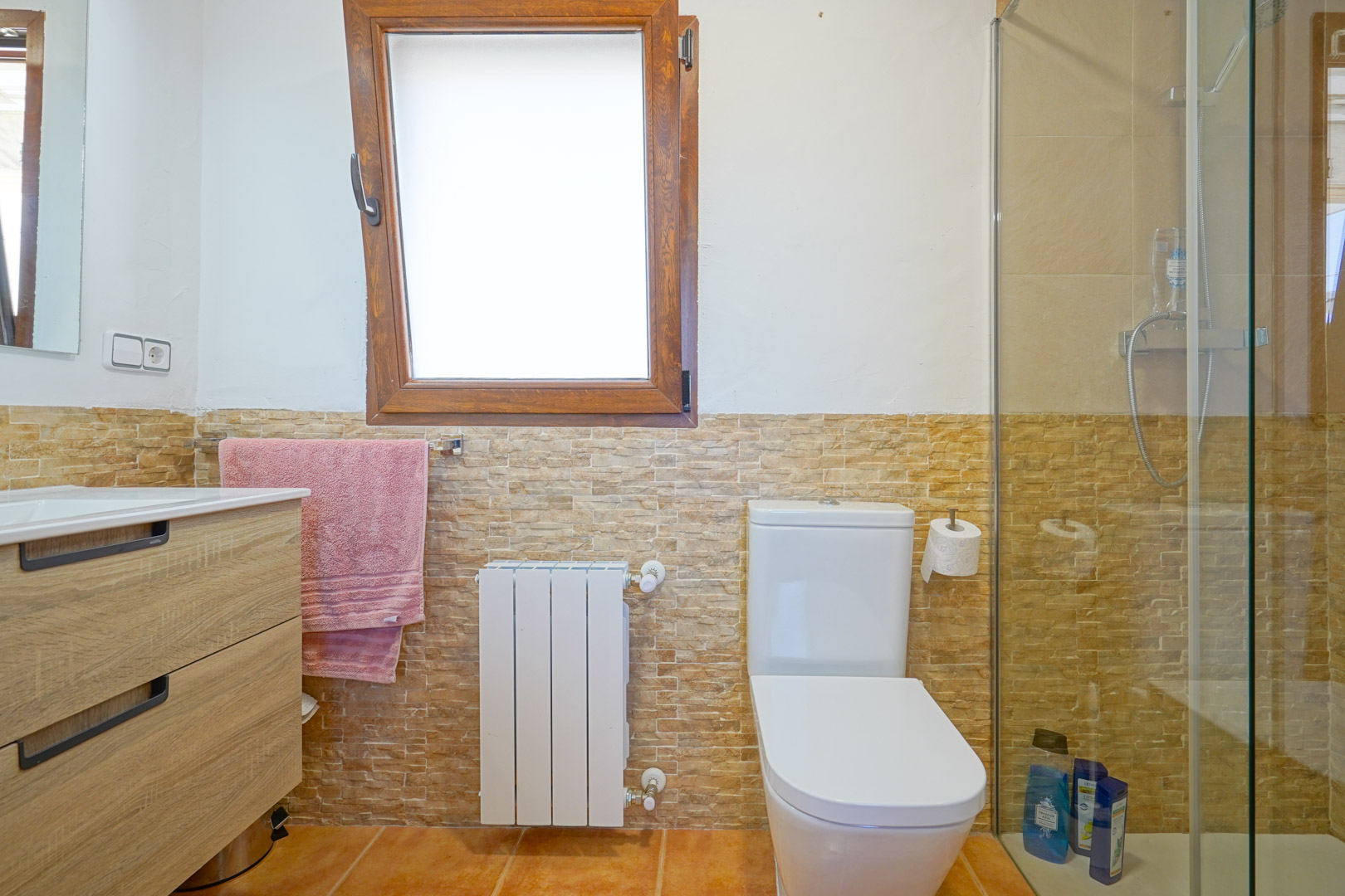 Badkamer met houten wastafelmeubel, wandtegels met steeneffect, een witte radiator en een inloopdouche met glazen wand naast een houten raamkozijn.