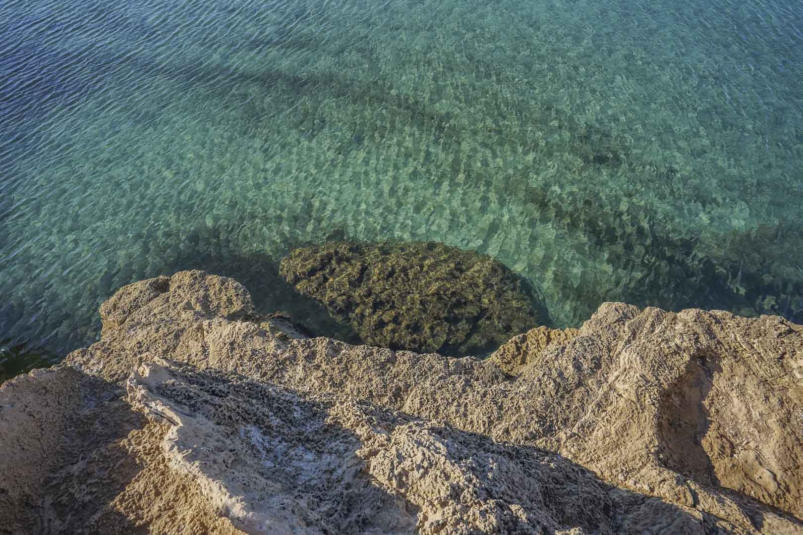 Bovenaanzicht van de rotsachtige kustlijn van Jávea, met kristalhelder turquoise water en kalksteenformaties zichtbaar onder het oppervlak van de Middellandse Zee.