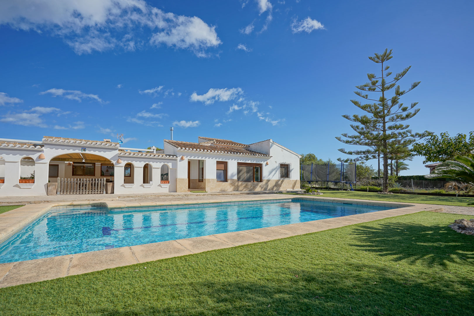 Buitenaanzicht van de villa met een rechthoekig zwembad, gazon en stenen terras. De witgestuukte gevel is voorzien van traditionele bogen en een mediterraan pannendak.
