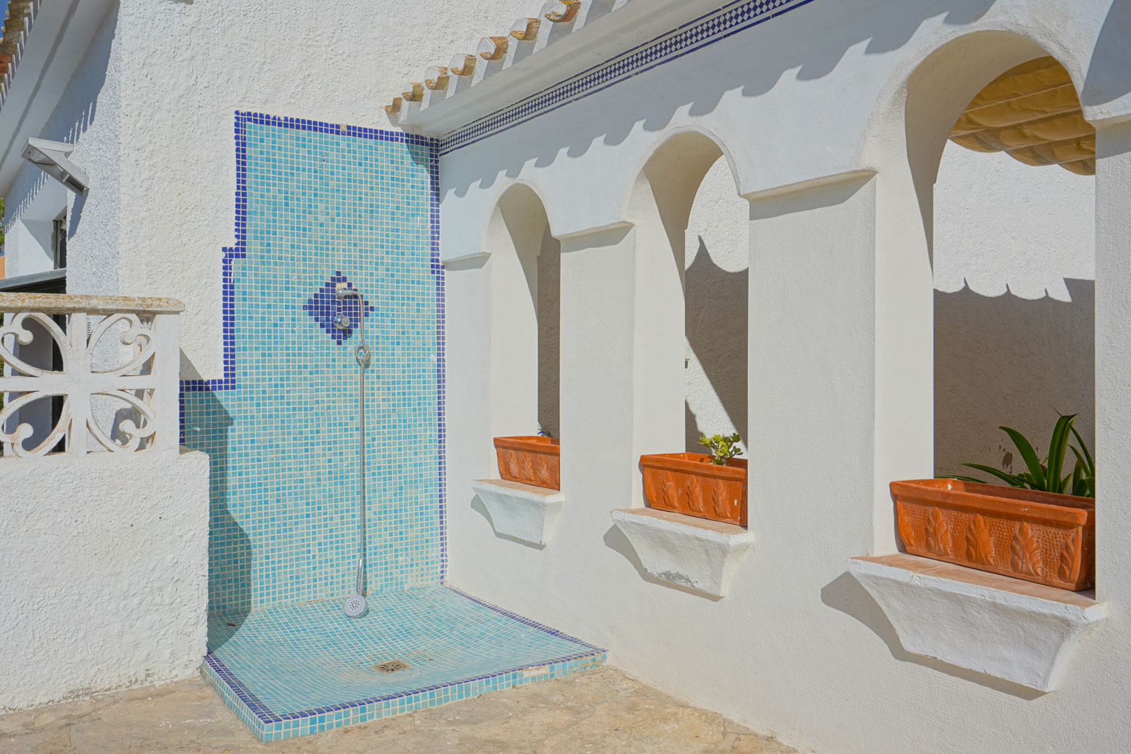 Buitendouche met blauwe mozaïektegels naast een witte gevel met mediterrane bogen en ingebouwde terracotta plantenbakken.