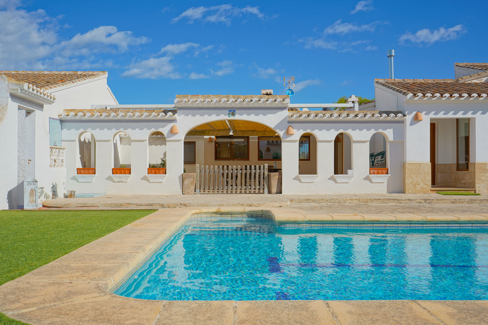 Buitenzwembad met natuurstenen rand en zonnig terras bij een witte mediterrane gevel met traditionele bogen en een dak van terracotta pannen.