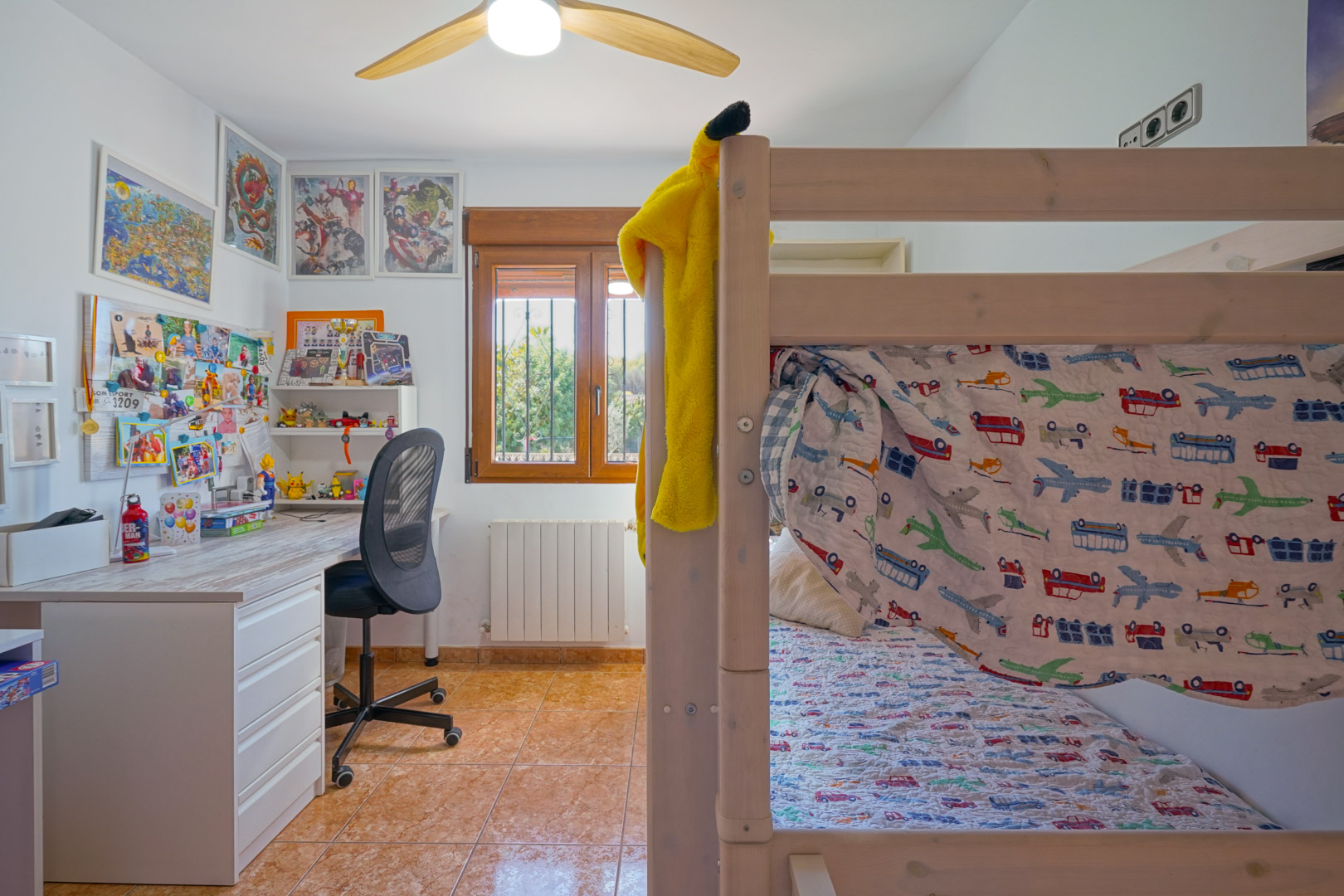 Kinderkamer met houten stapelbed, bureau en raam met houten kozijnen. De kamer is voorzien van een radiator, plafondventilator en een tegelvloer.