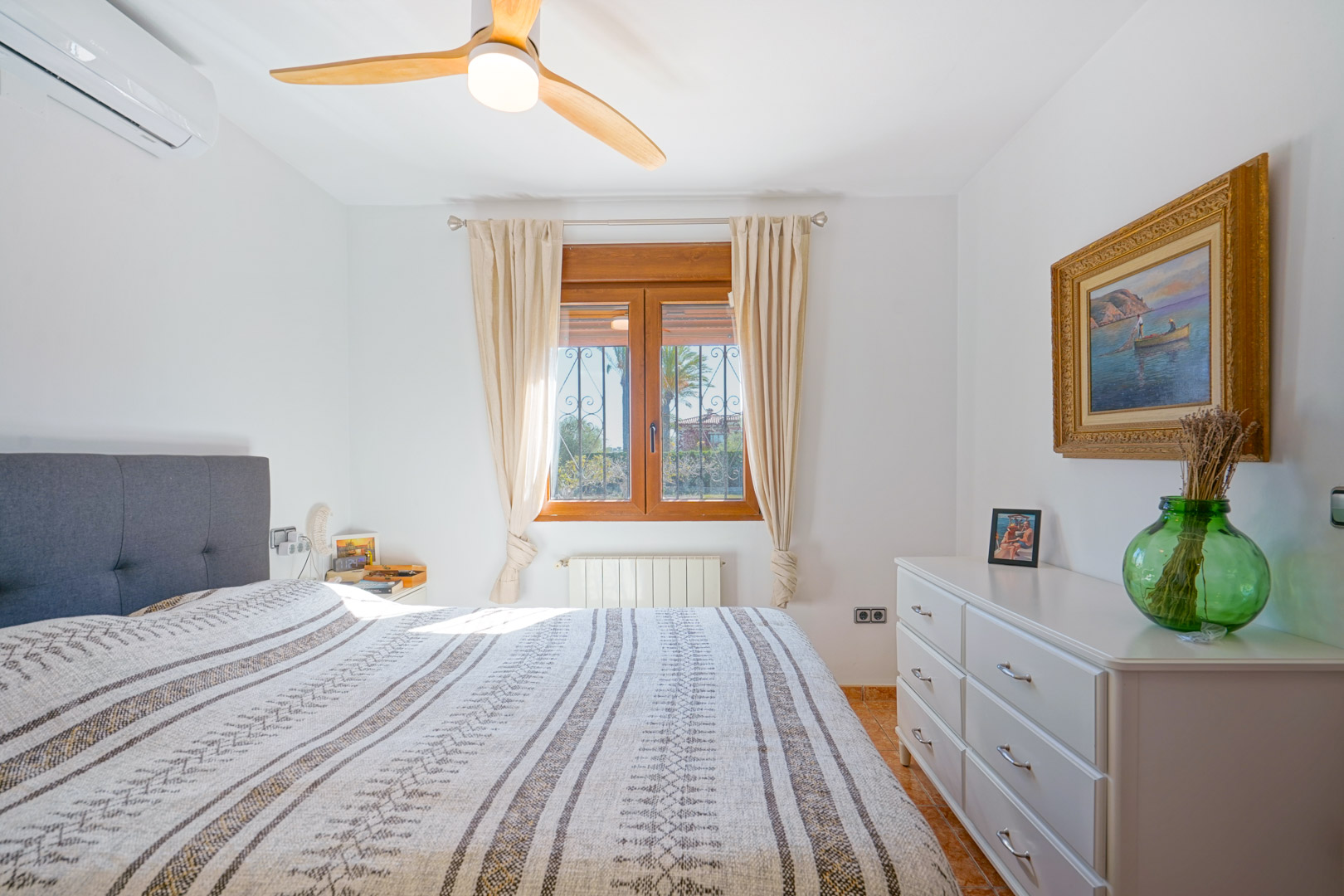 Lichte slaapkamer met houten kozijn, plafondventilator met houten bladen, airconditioning en een witte commode. Voorzien van radiatorverwarming en een tegelvloer.