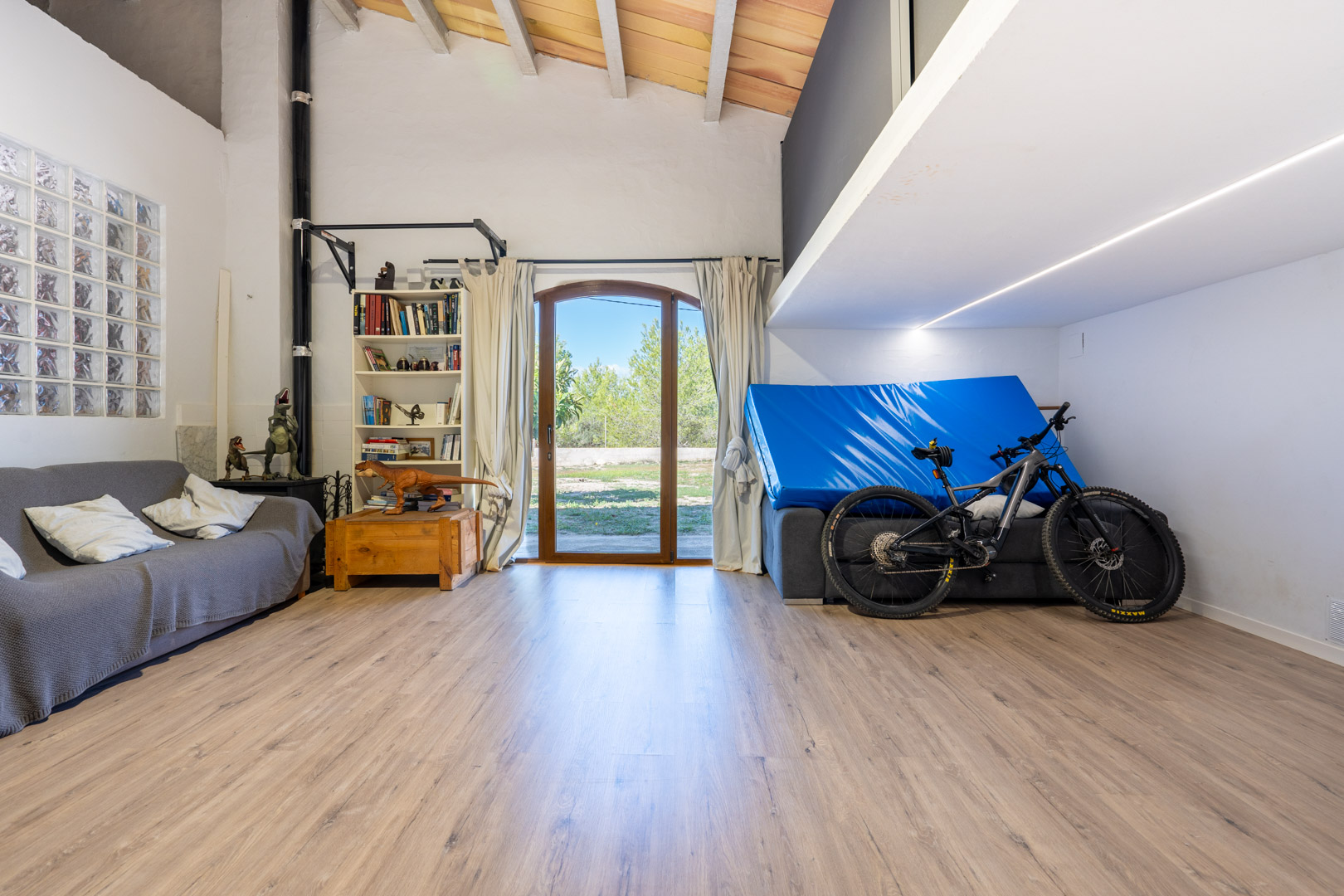 Multifunctionele ruimte met hoge plafonds met houten balken, een entresol, laminaatvloer en een boogdeur die direct toegang biedt tot de tuin.
