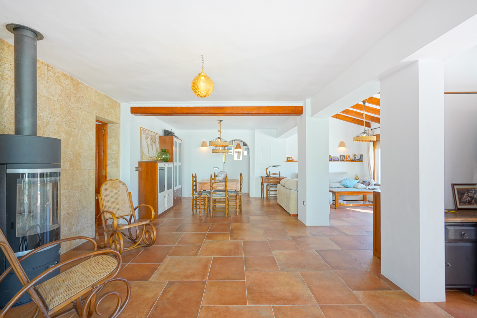 Open woonkamer met terracotta vloertegels, een moderne houtkachel tegen een natuurstenen wand en rustieke houten balken die de eethoek markeren.