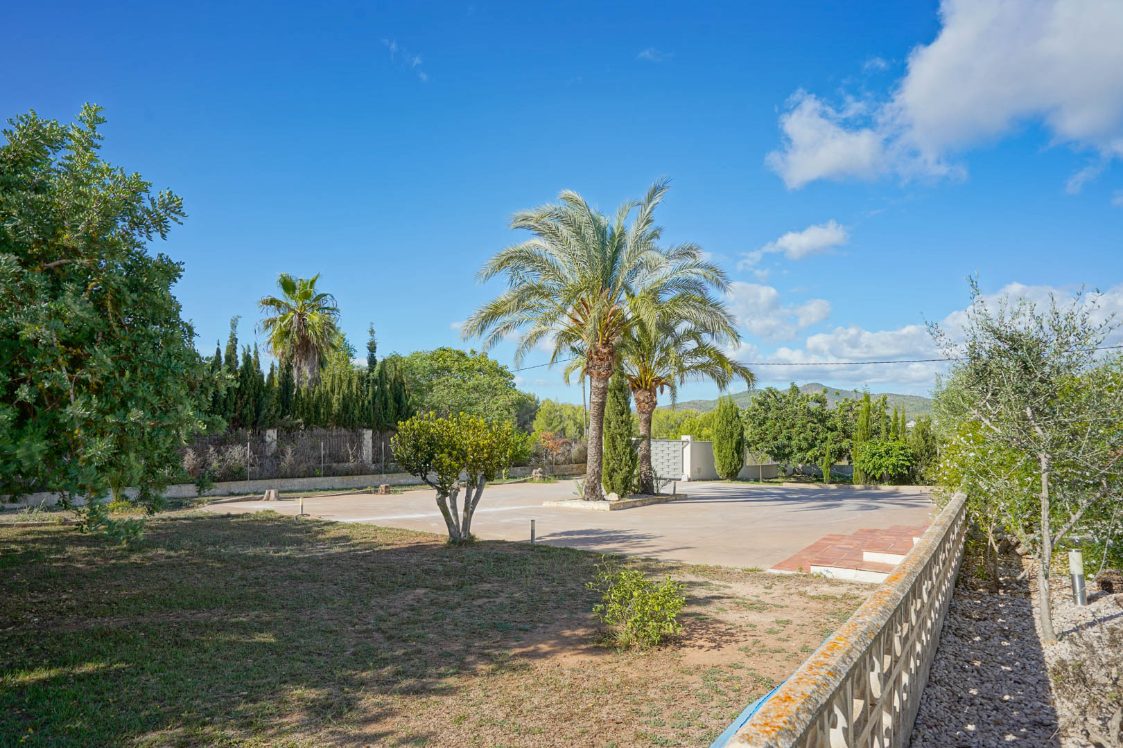 Royale geplaveide oprit met centrale palmbomen, omgeven door mediterrane beplanting en uitzicht op de bergen in de verte onder een blauwe hemel.