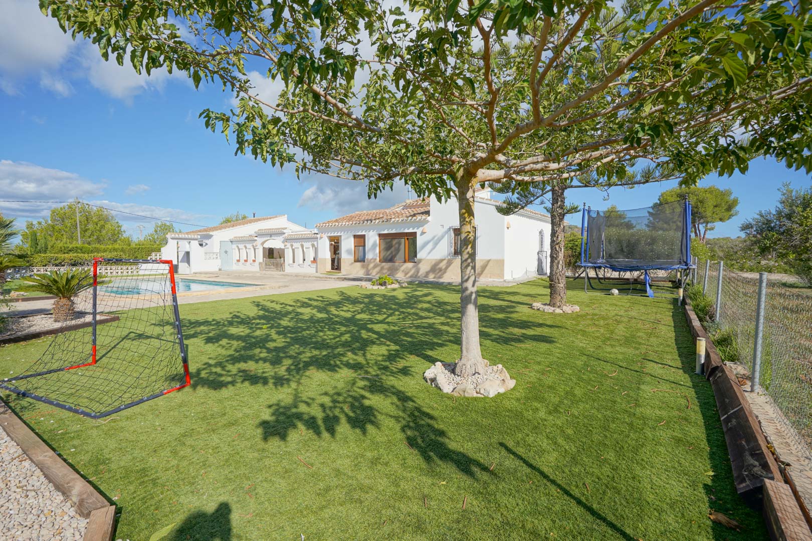 Royale tuin met kunstgras en recreatieruimte voorzien van een voetbalgoal en trampoline, grenzend aan de witte gevel van de villa en het zwembad.