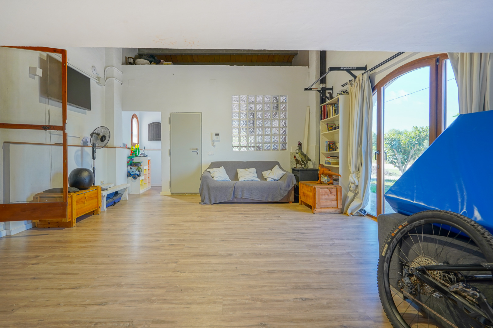 Ruime multifunctionele kamer met laminaatvloer, zithoek en veel lichtinval via glasblokken en grote openslaande deuren naar de tuin.