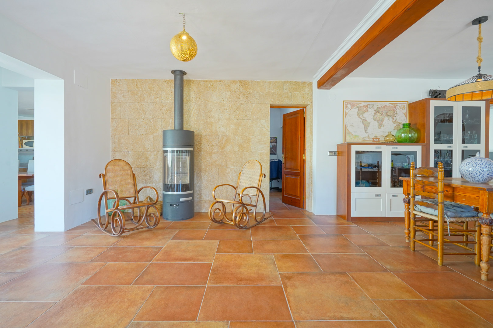Woon-eetkamer met terracotta vloertegels, een moderne houtkachel tegen een natuurstenen muur en rustieke houten plafondbalken.