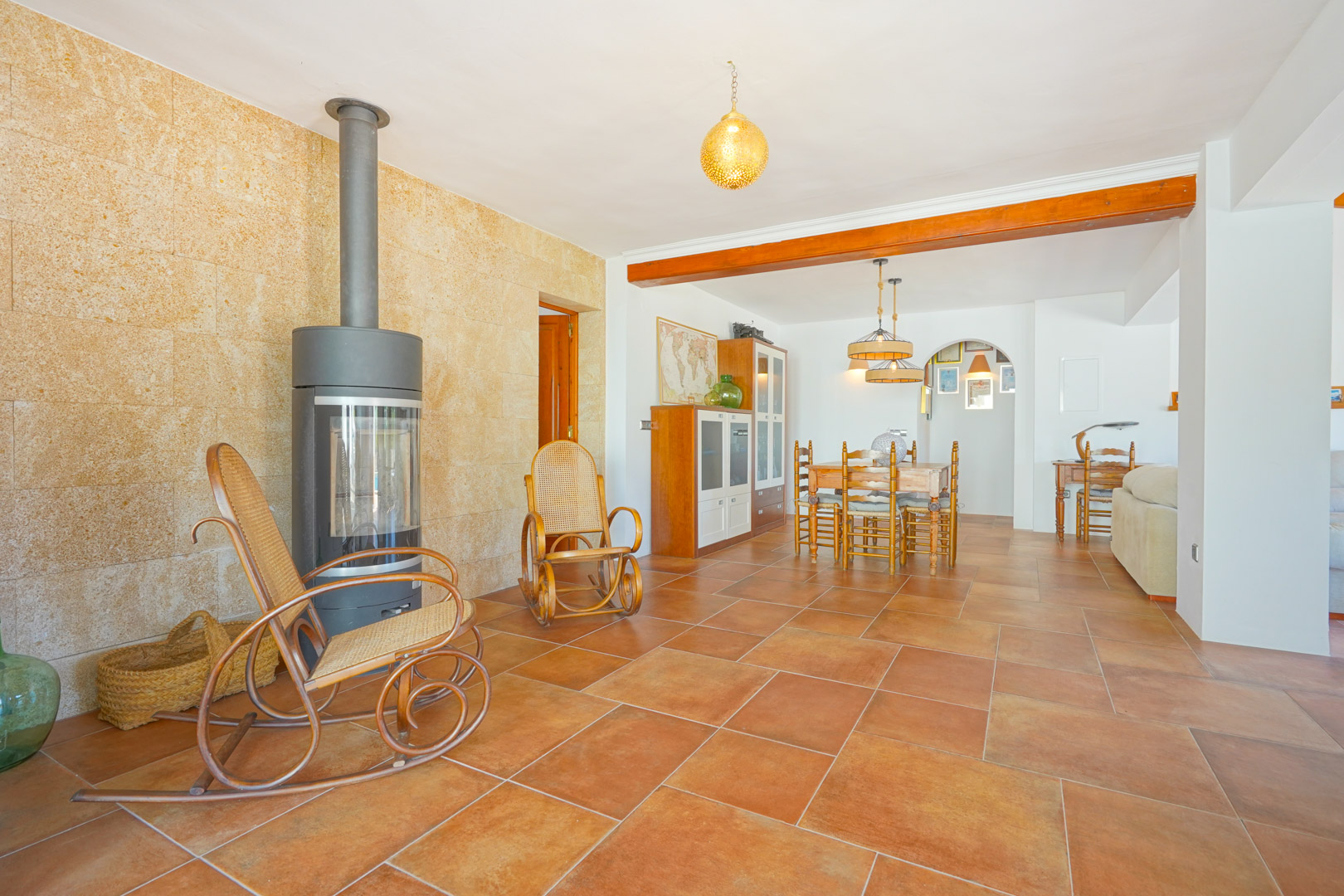 Woonkamer met een moderne houtkachel tegen een natuurstenen wand, terracotta vloertegels en een eethoek onder een rustieke houten balk.