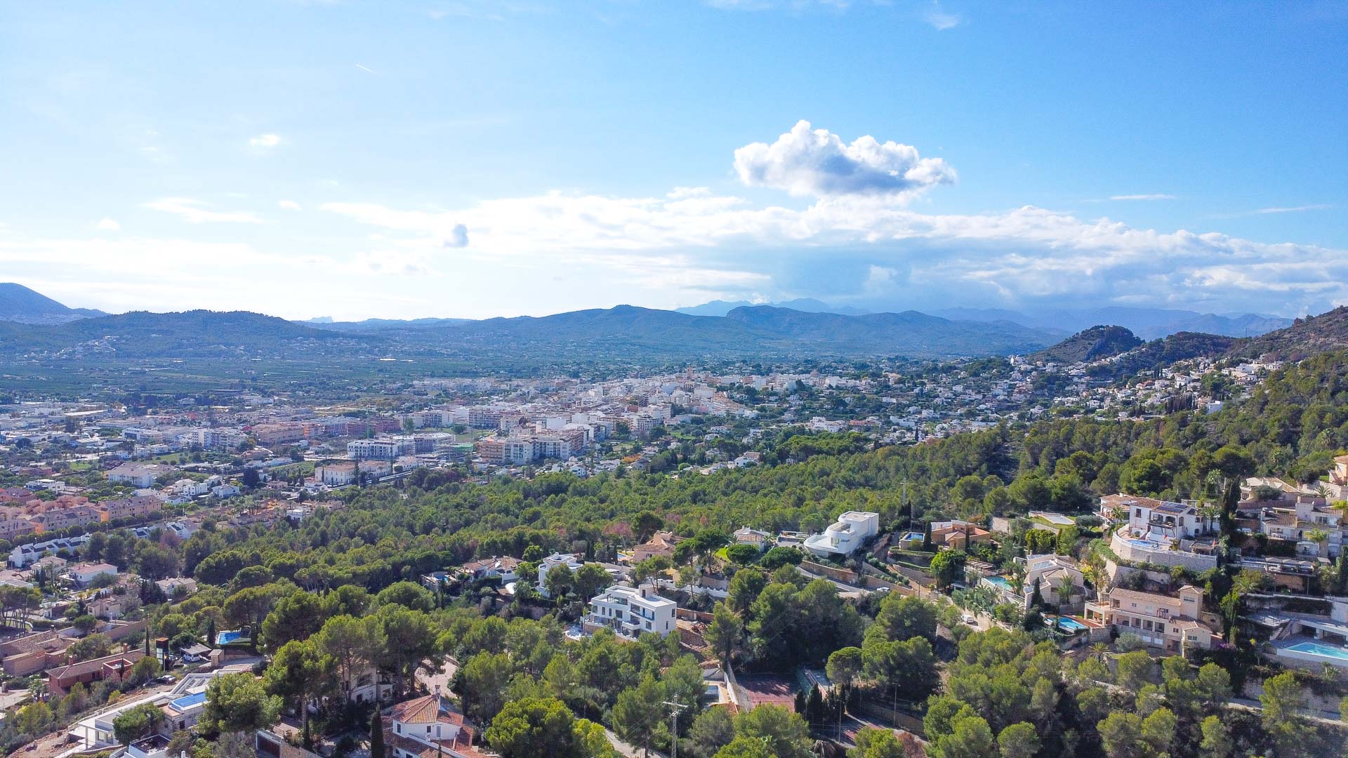 Luchtopname van de woonwijk La Corona in Jávea, met focus op de bosrijke heuvels, de verspreide villa's en het weidse uitzicht over de vallei en bergen.