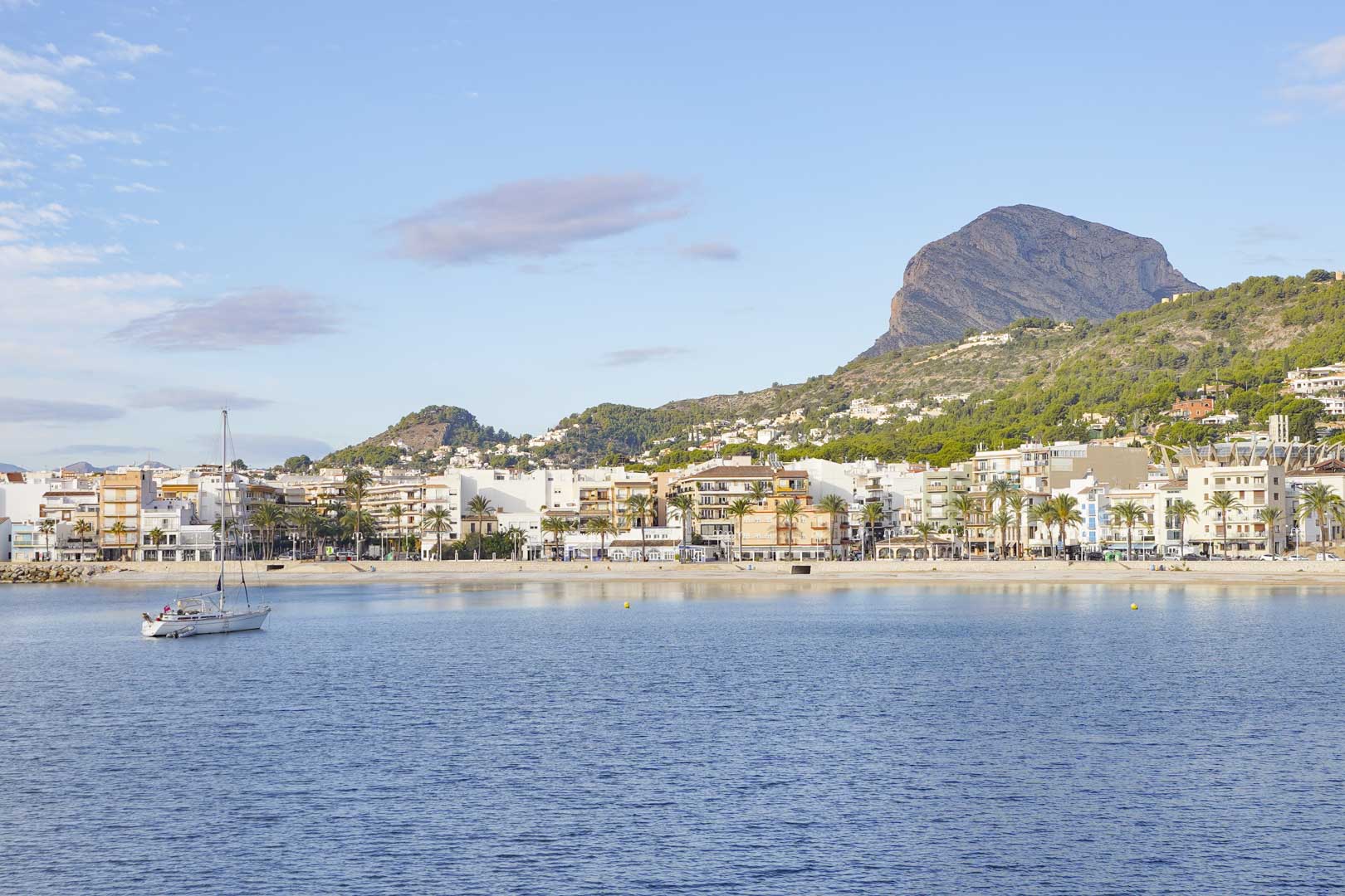 Panoramisch uitzicht op de haven en het strand van Jávea met de Montgó op de achtergrond. Een zeilboot ligt in het rustige water voor de met palmen omzoomde boulevard.