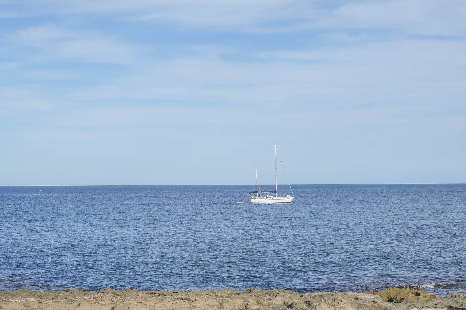 Panoramisch uitzicht op de Middellandse Zee vanaf de kust van Jávea, met een zeilboot voor de rotsachtige kustlijn onder een heldere hemel.